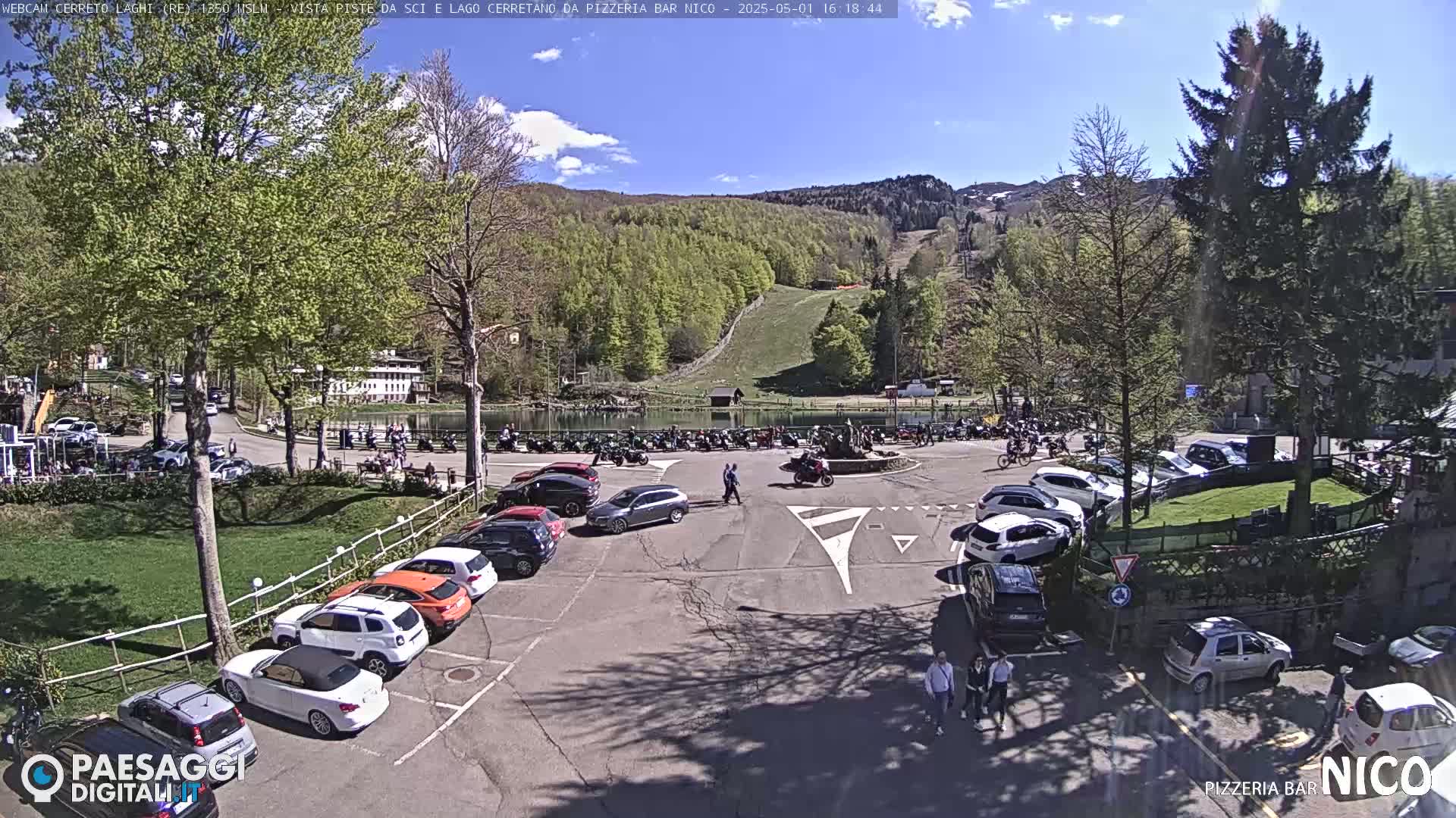 A sunny day shows a parking lot full of cars near a lake and a mountainside with a ski slope, surrounded by green trees.