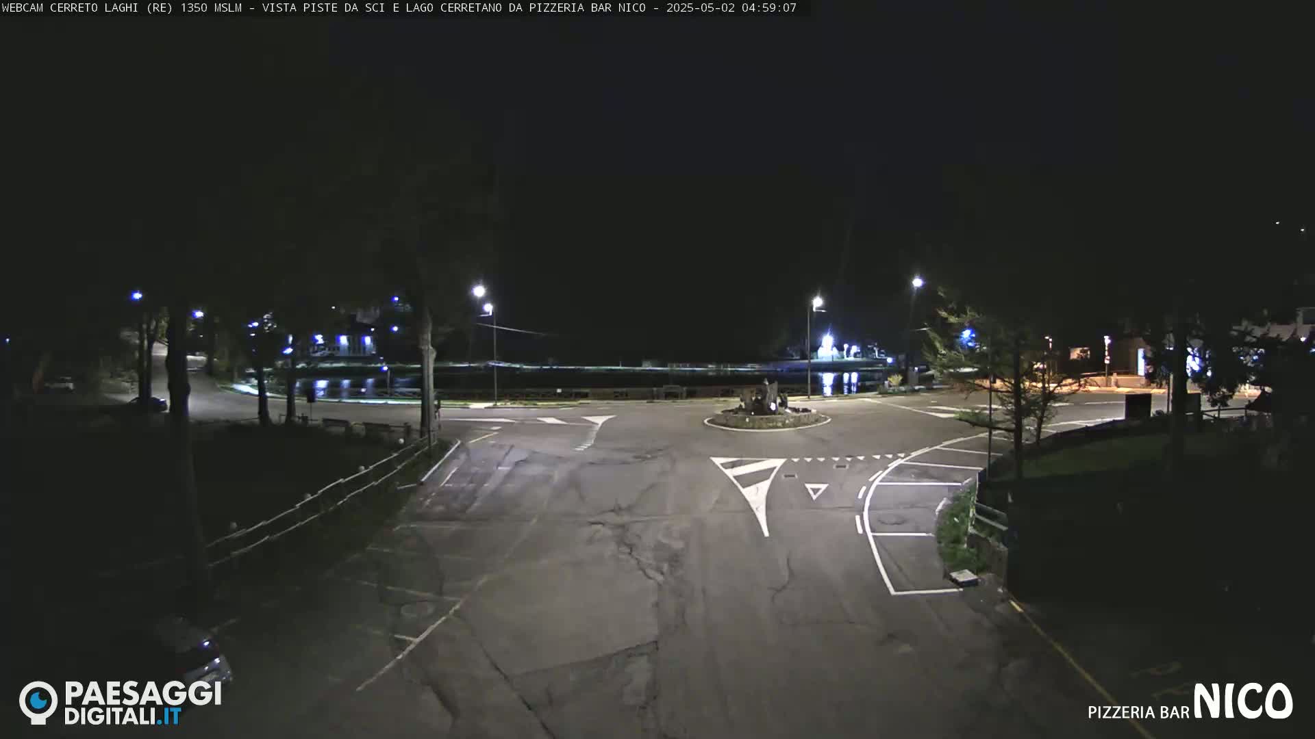 A nighttime, dimly lit view of an empty roundabout and surrounding parking area near a body of water.