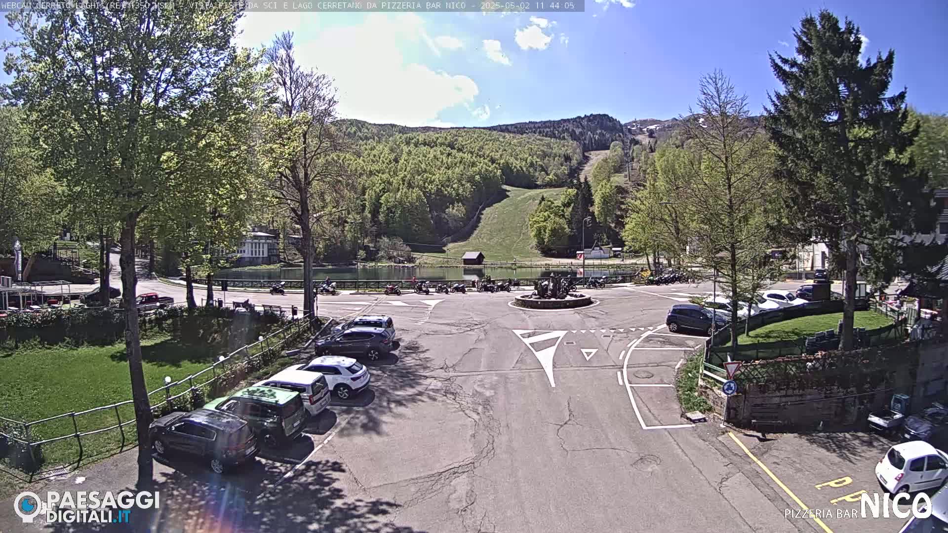 A sunny day reveals a parking lot, a small pond, and a hillside with greenery and a ski slope visible in the background.
