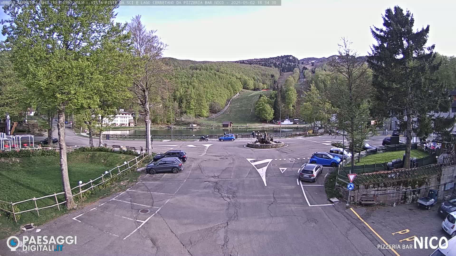 A parking lot with several cars is situated near a small lake and a hillside with green trees under a partly sunny sky.