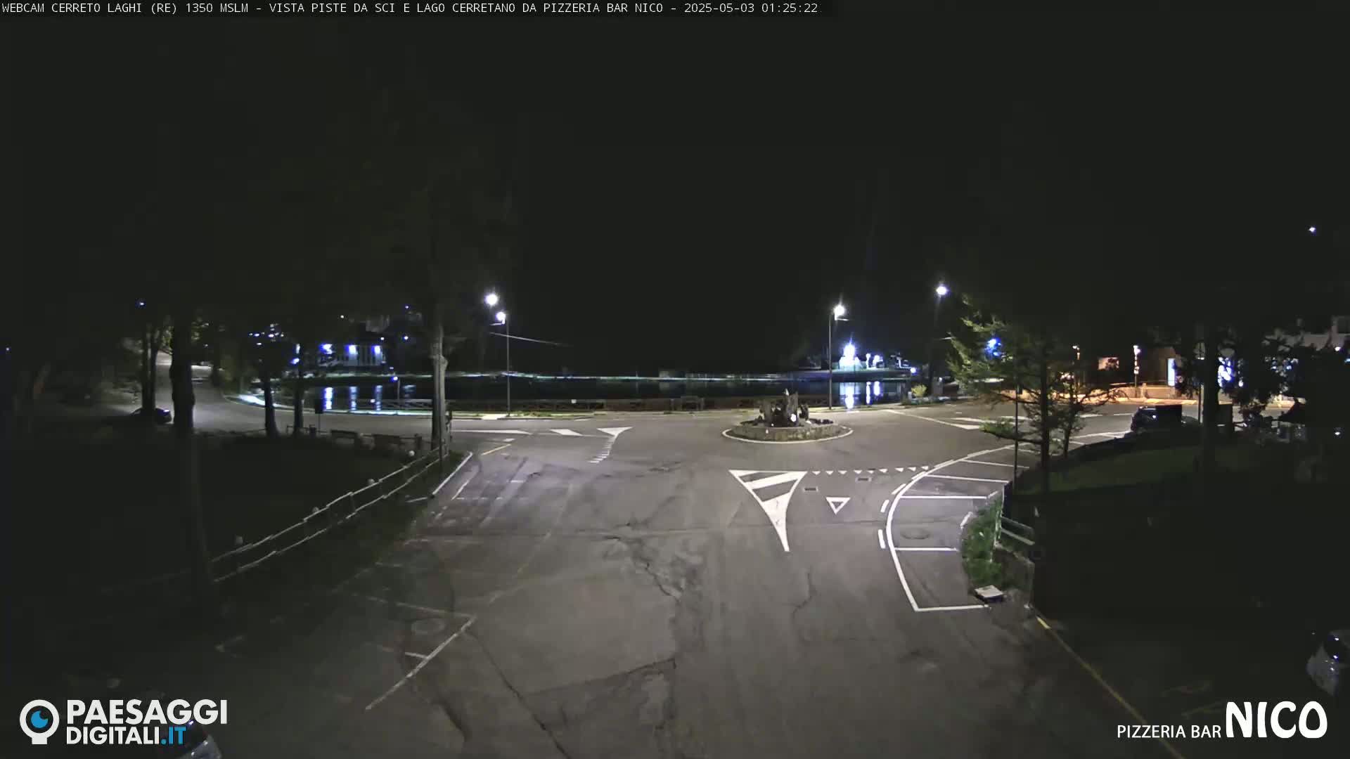 A nighttime view of a dark, empty roundabout near a body of water, illuminated by streetlights.