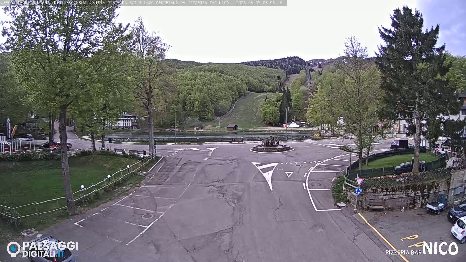 A mostly empty parking lot and roundabout are situated in front of a mountain with green trees and ski slopes under a cloudy sky.