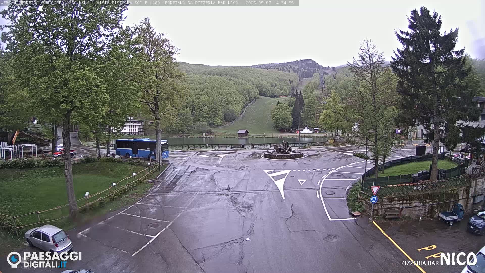 A wet parking lot and roundabout are situated in front of a small town nestled in a valley of green hills and trees, under an overcast sky.