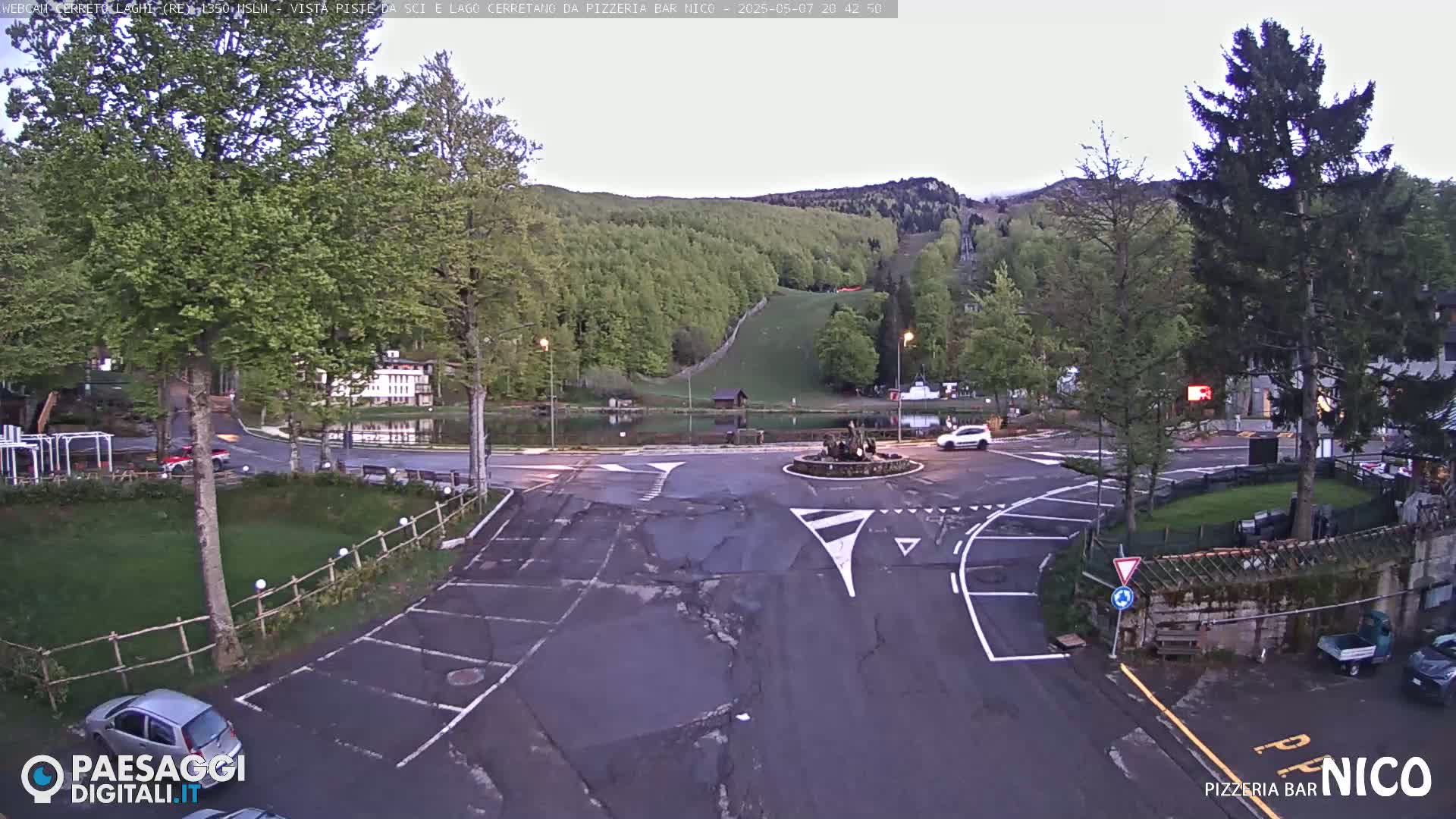 A parking lot and roundabout are seen near a small lake, with green hills and a ski slope in the background under a mostly clear, twilight sky.