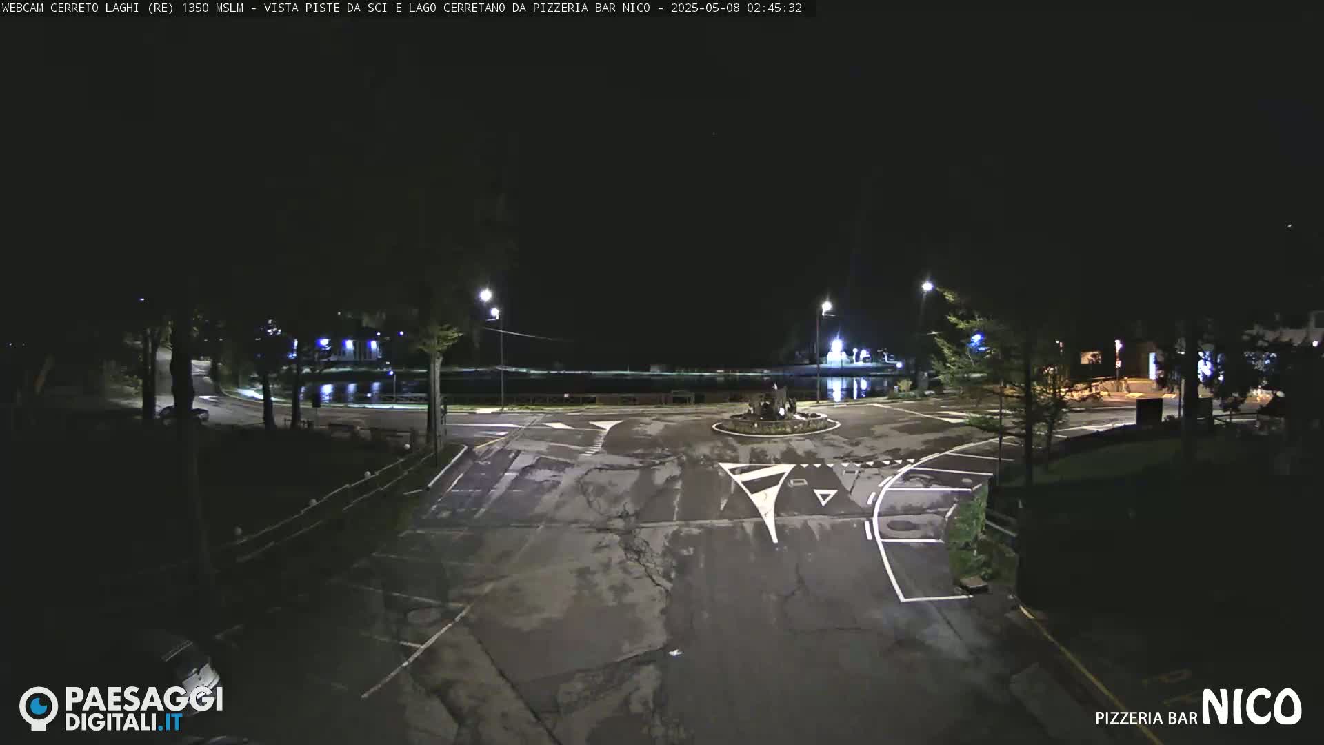 A nighttime view of an empty parking lot and roundabout near a body of water, illuminated by streetlights.