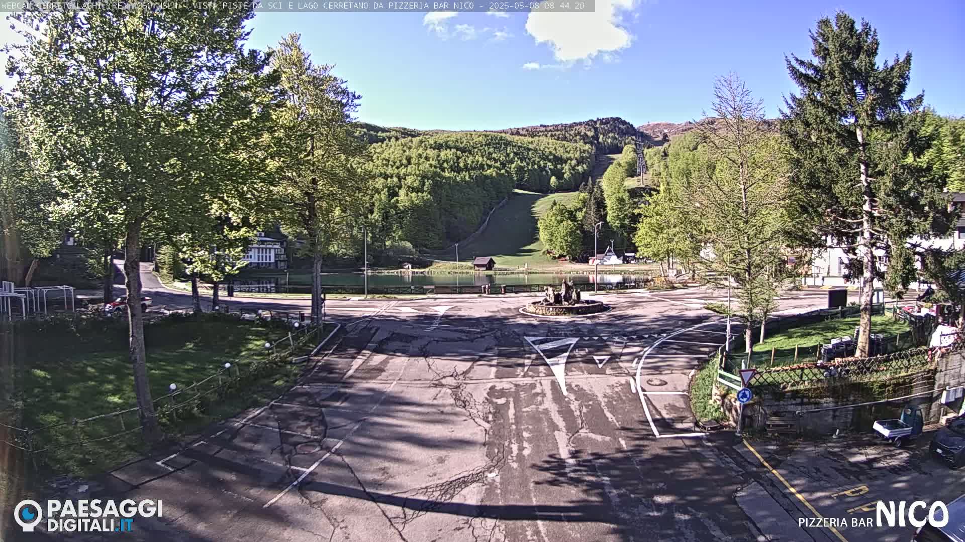 A sunny view of a paved area with a fountain in the center, surrounded by green trees, a small pond, and a ski slope in the background.