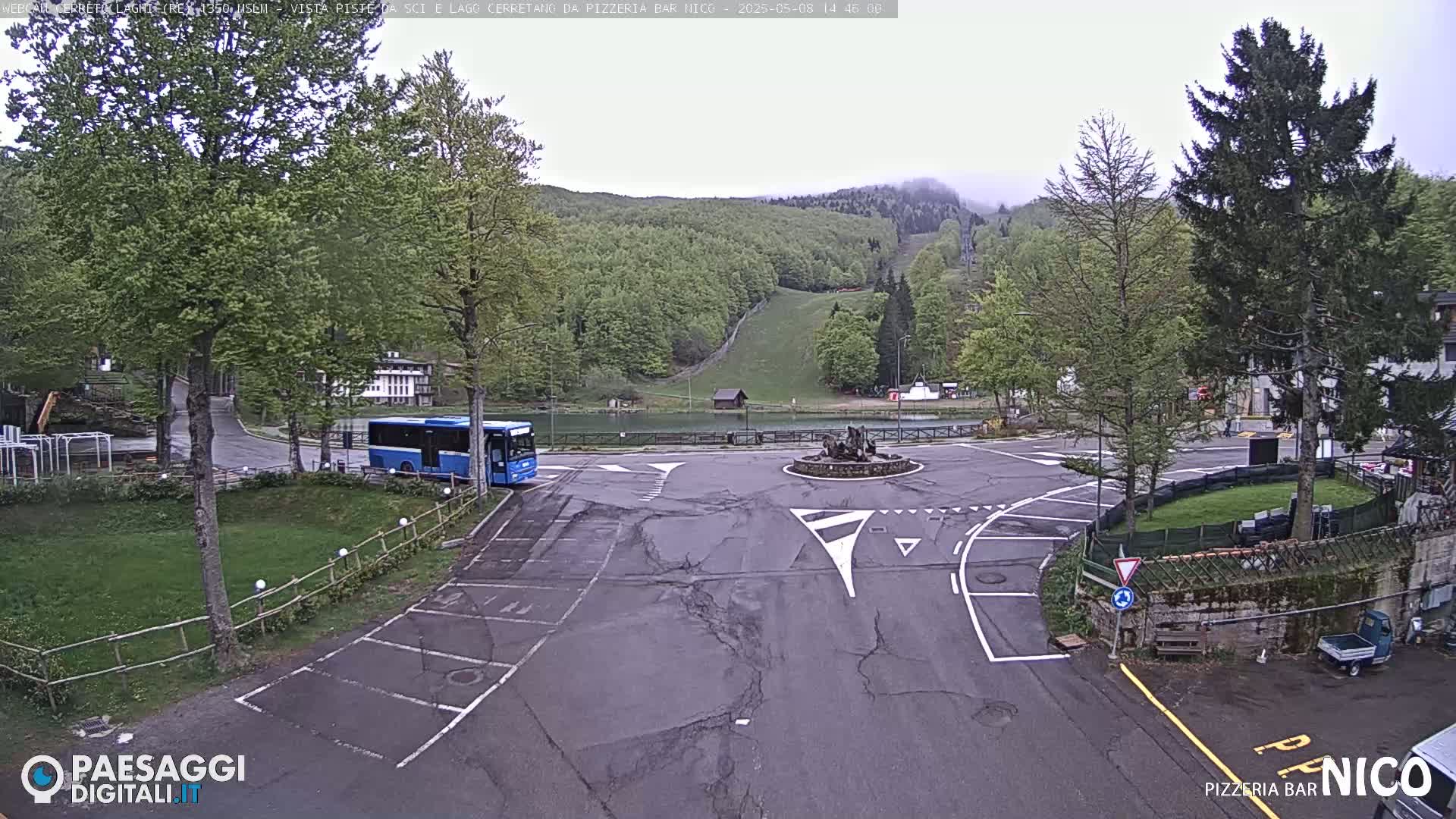 A mostly empty roundabout with a blue bus parked nearby, situated in front of a wooded hillside with a ski slope visible under an overcast sky.