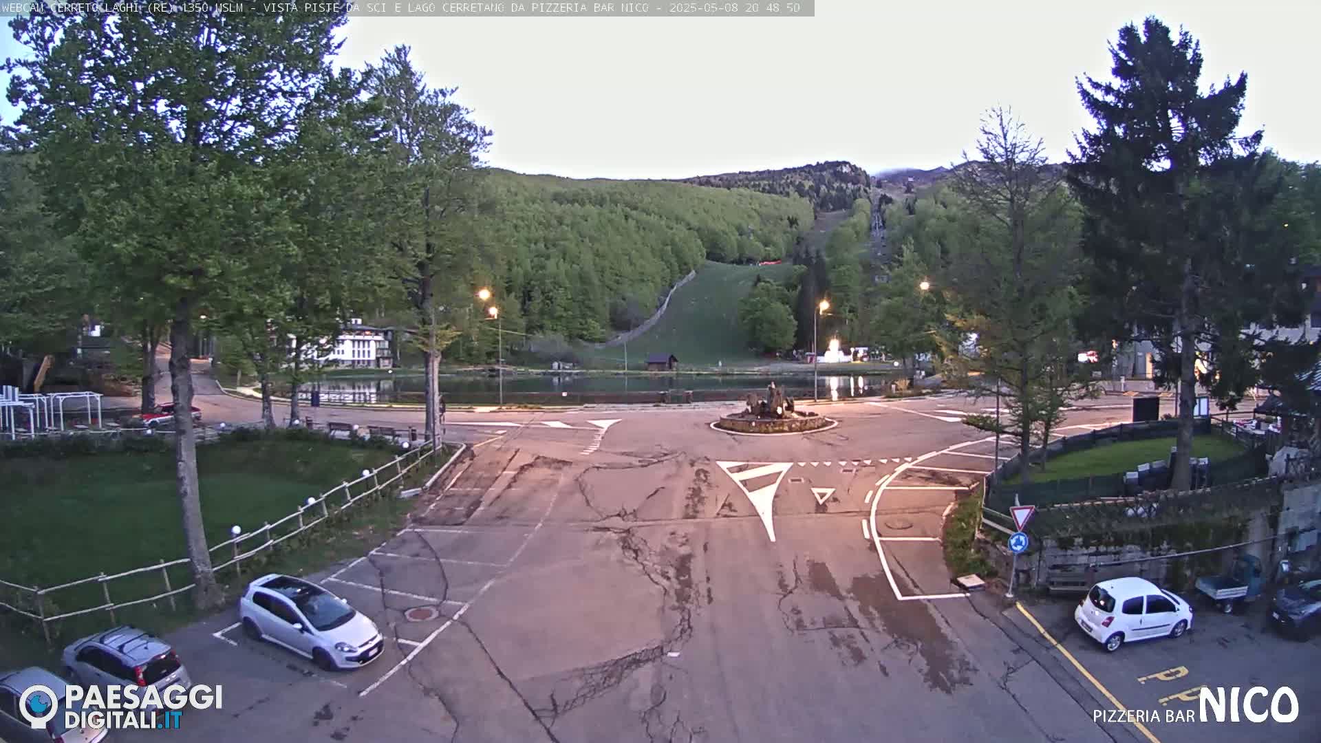 A twilight view of a roundabout with a small pond and fountain, surrounded by trees, buildings, and parked cars, overlooking a grassy hill with ski slopes in the background.