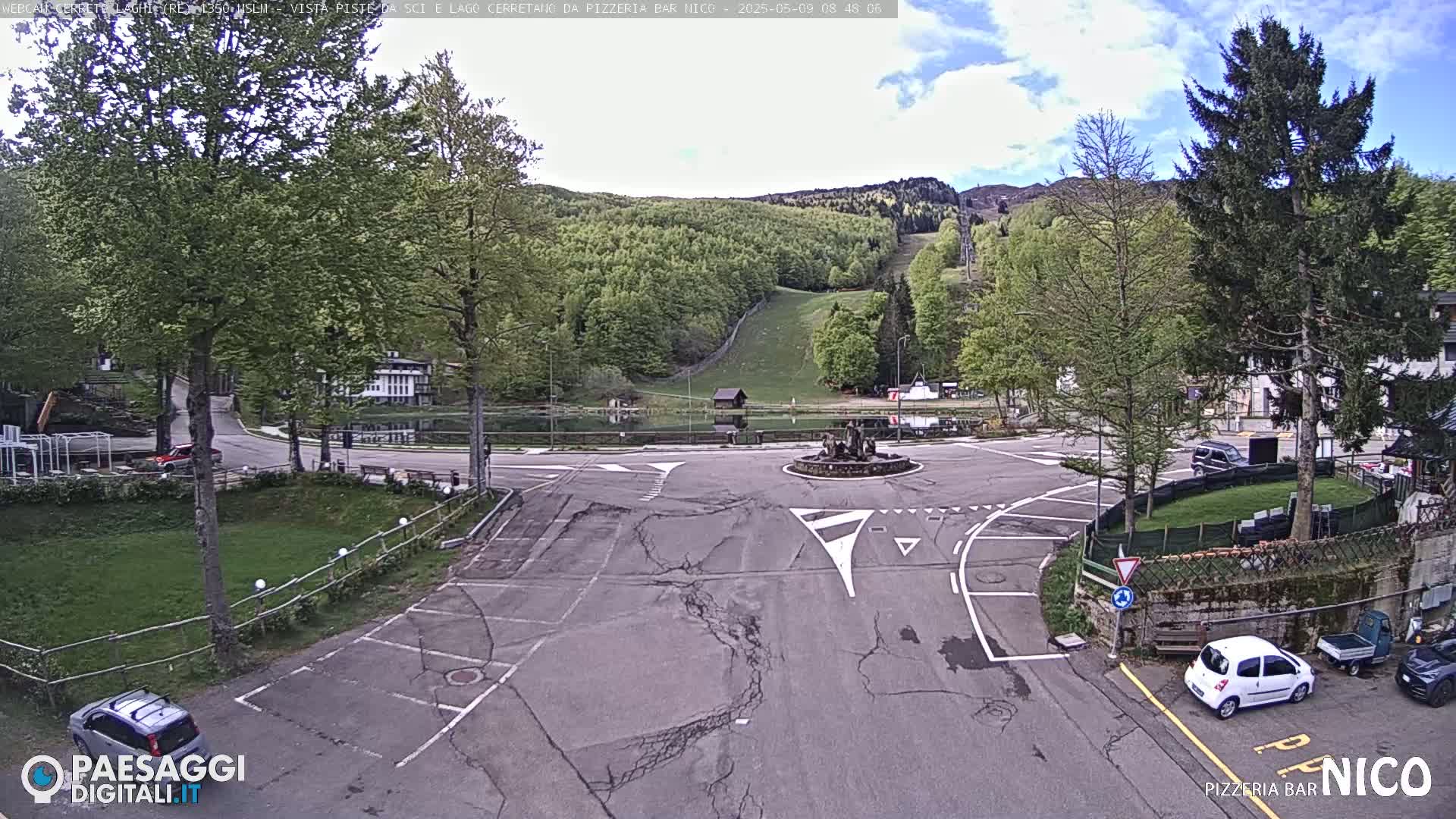 A mostly sunny day shows a paved roundabout with a fountain at its center, surrounded by green grass, trees, buildings, and parked cars, with a hillside and ski slope visible in the background.