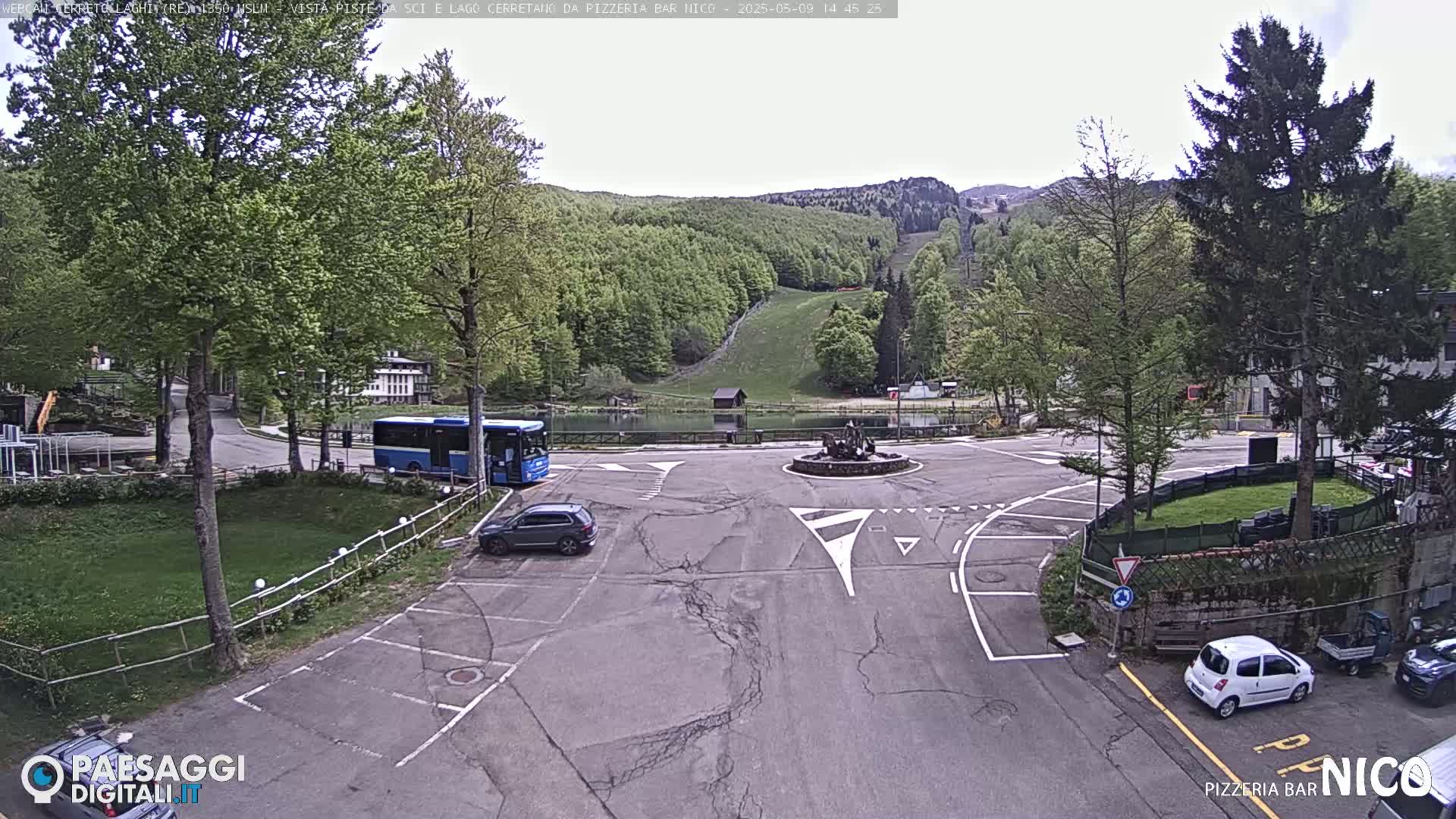 A parking lot with a few cars and a bus, situated near a small pond and a forested hillside under an overcast sky.