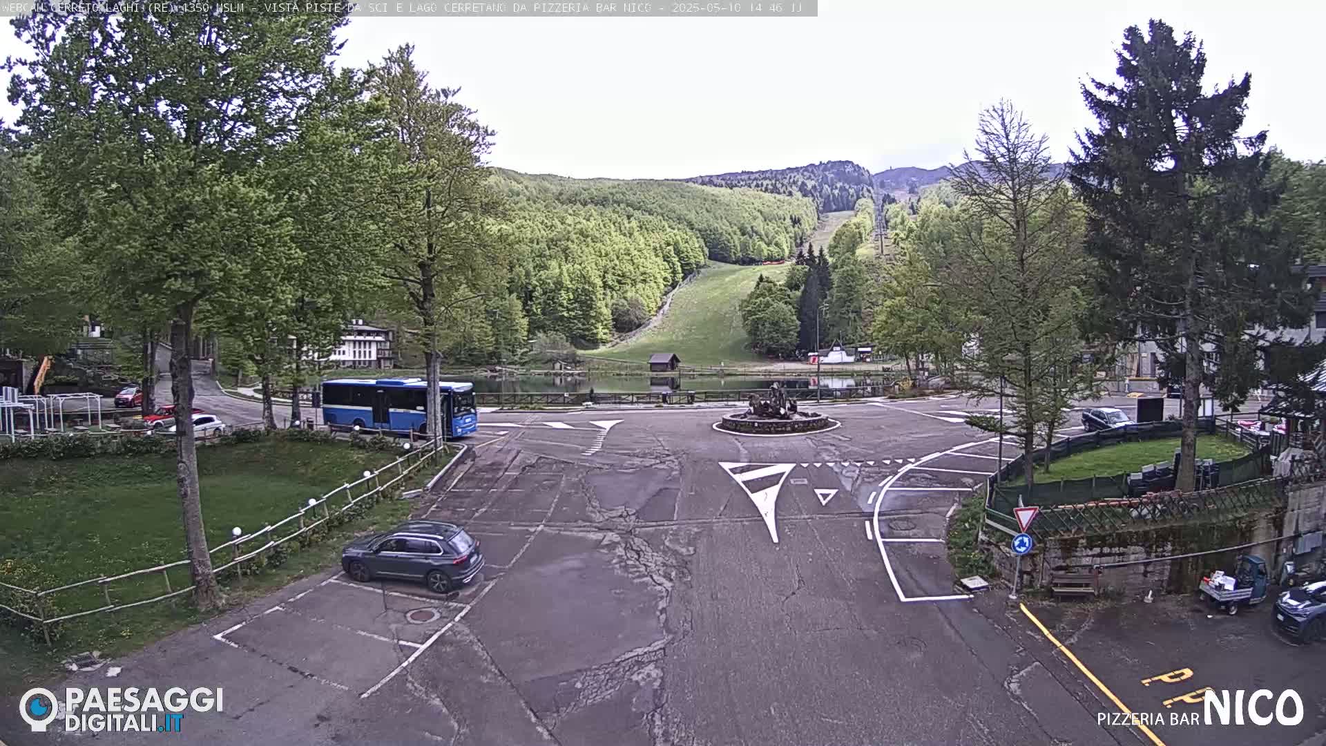 A parking lot and roundabout are shown in an overcast setting, with a bus, cars, and a small pond visible, surrounded by green trees and a hill with a ski slope in the background.
