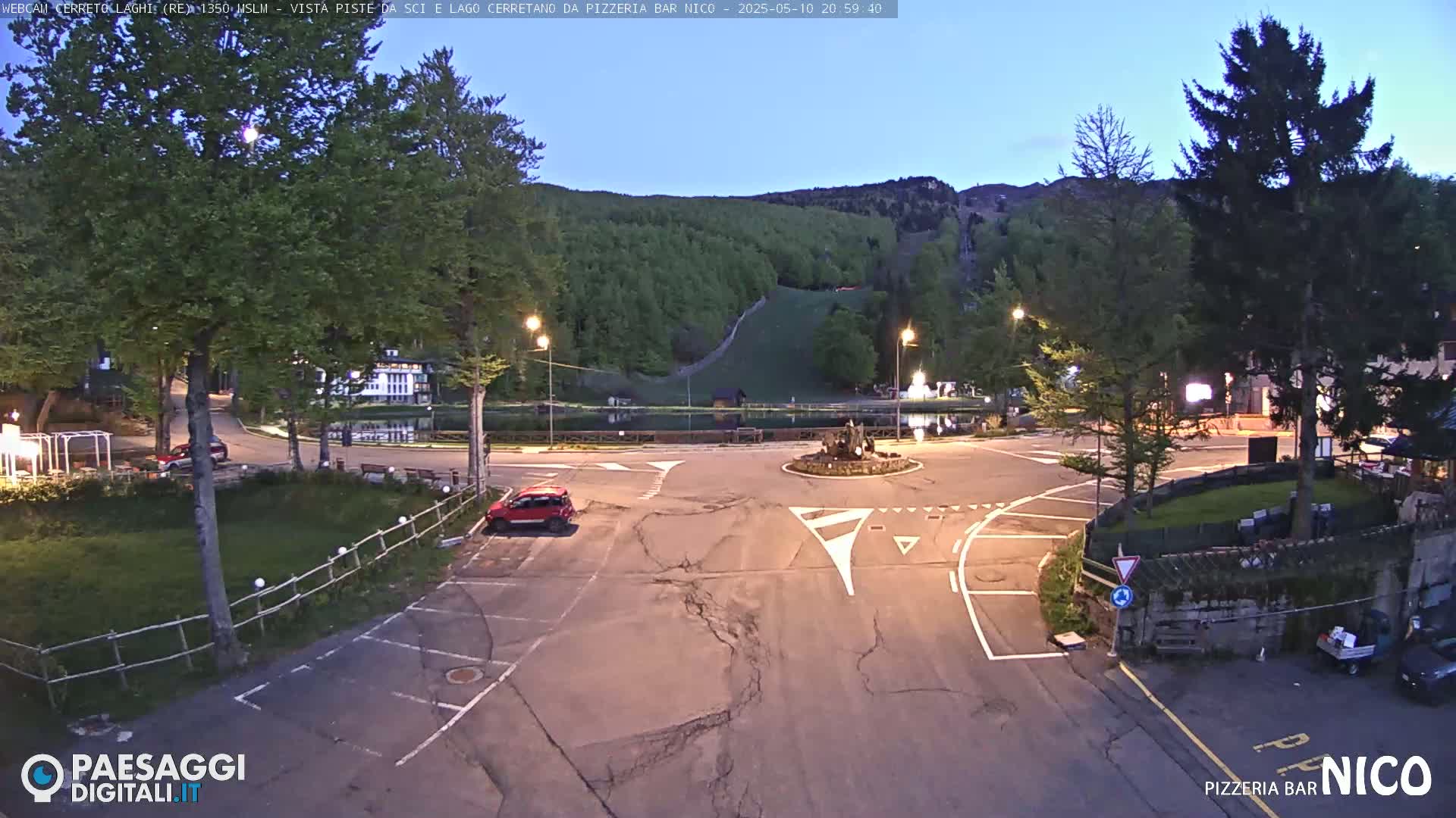 A twilight view of a town square with a small pond, surrounded by trees and buildings, a parking lot, and a few cars.