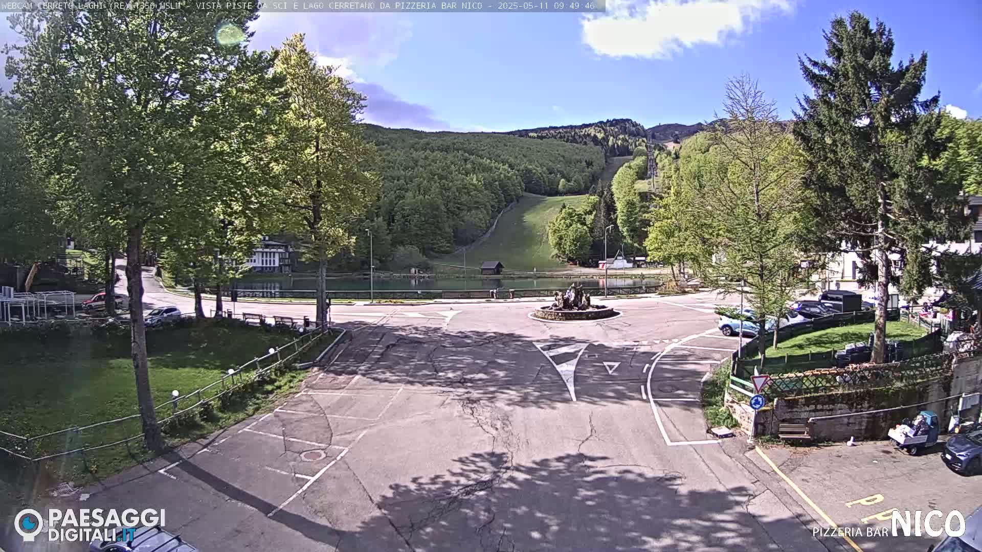 A sunny day reveals a roundabout in a town next to a lake and forested hillside with a ski slope visible in the distance.