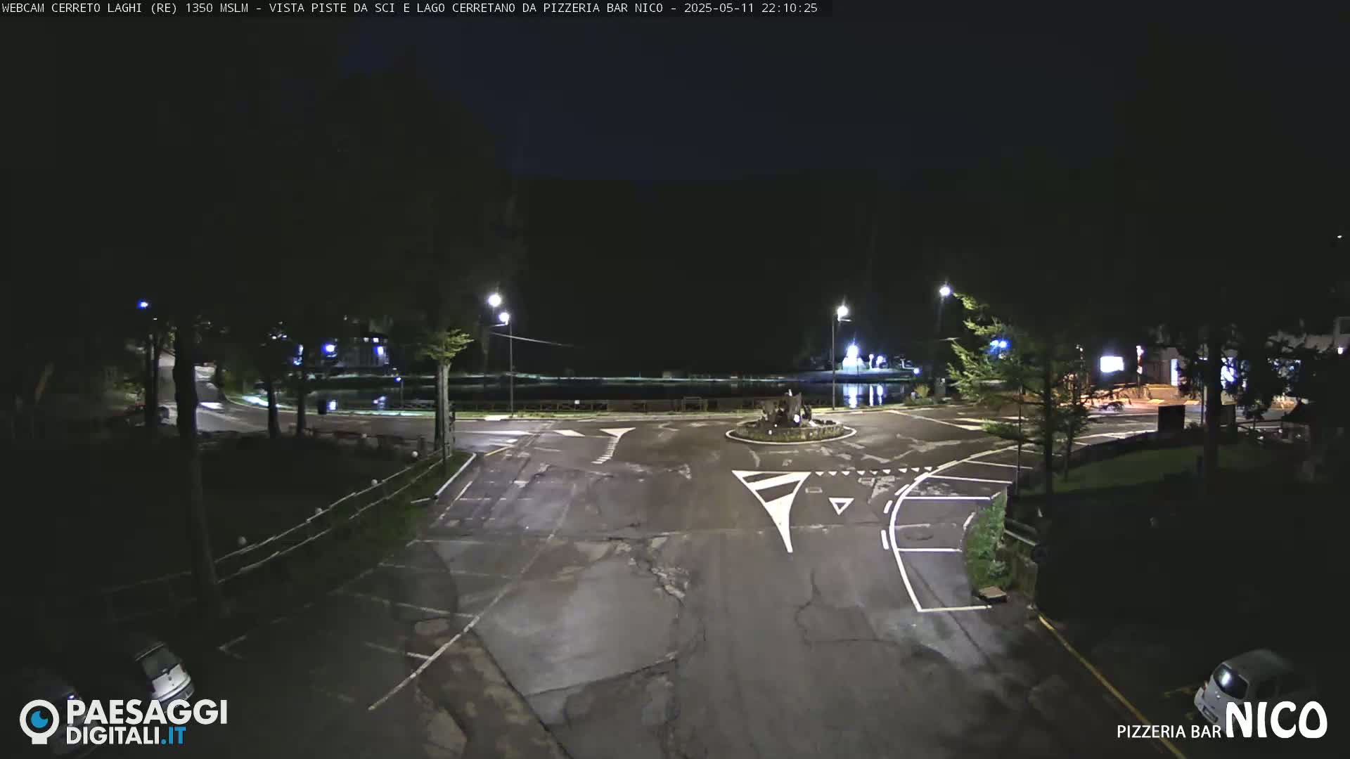 A nighttime, empty parking lot and roundabout are illuminated by streetlights near a body of water.
