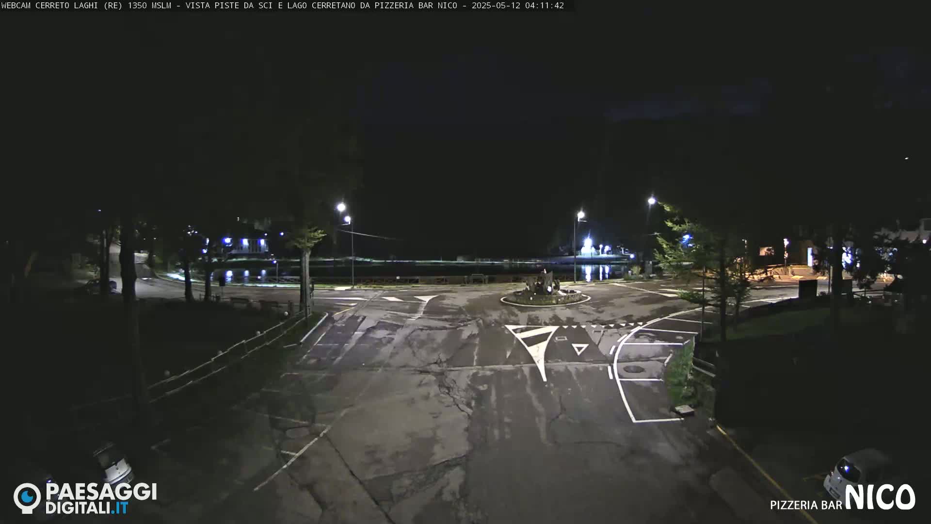 A nighttime view of an empty parking lot and roundabout near a body of water, illuminated by streetlights.