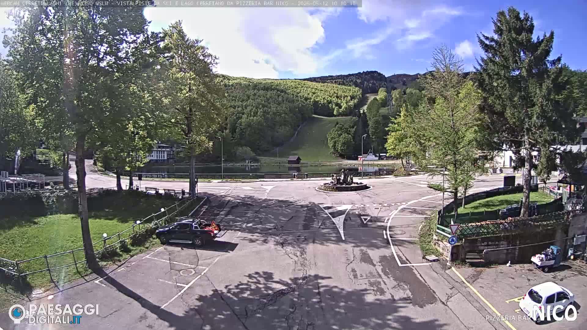 A sunny view of a parking lot and roundabout near a small lake and ski slope, surrounded by green trees and mountains.