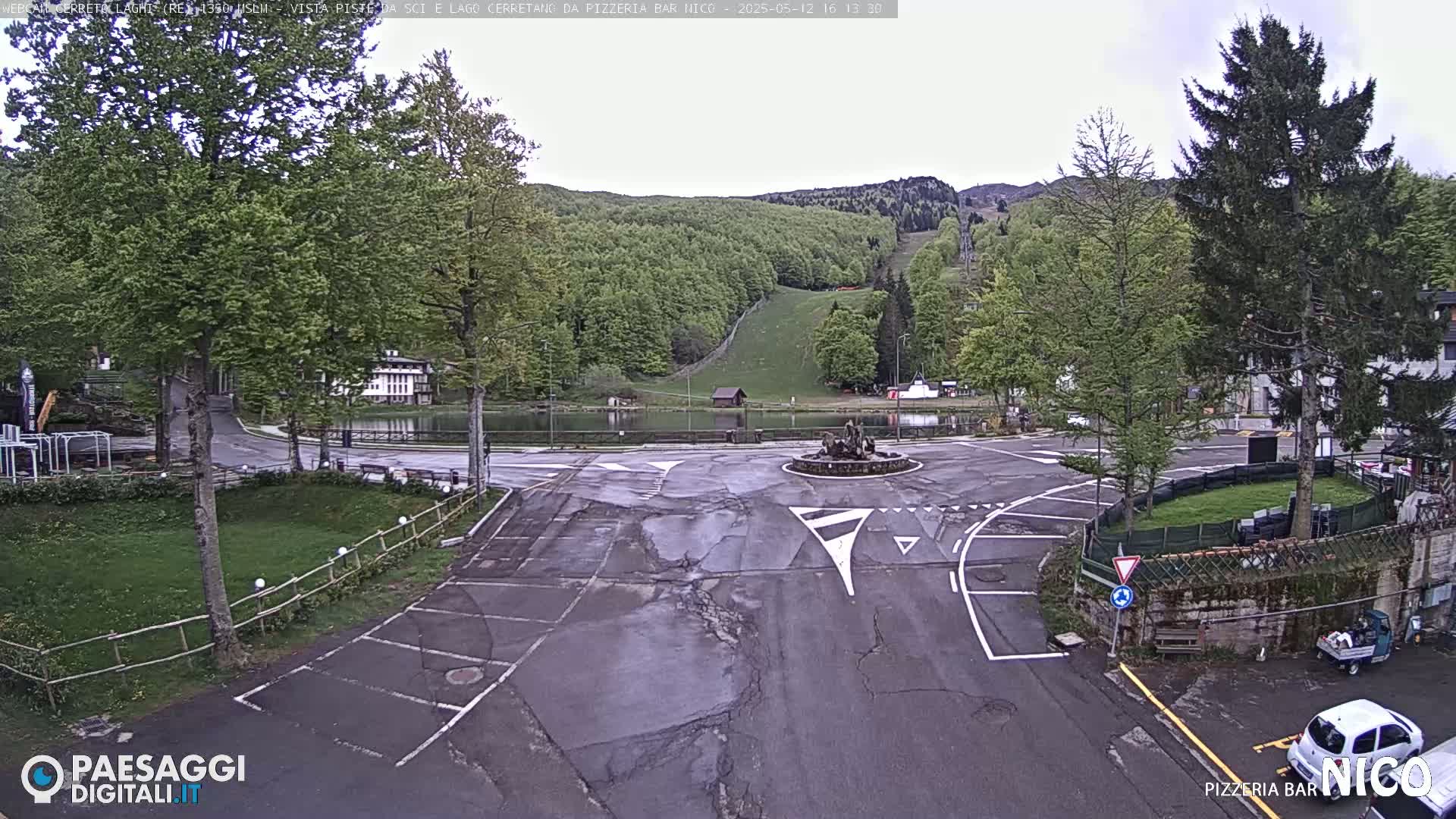 A wet, paved area with parking spaces and a small pond, surrounded by green trees and a hillside with a ski slope in the background under an overcast sky.