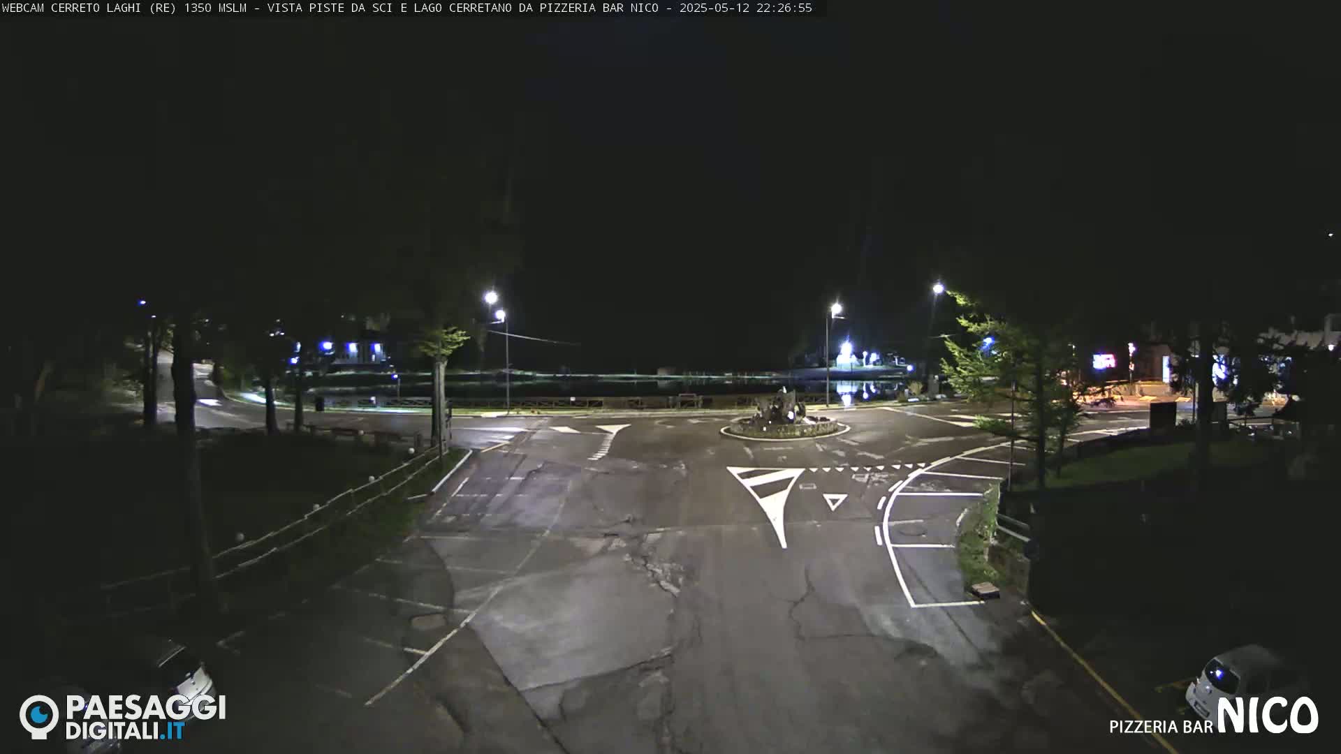 A nighttime view of a mostly empty roundabout and parking lot with a few cars visible under dark, clear skies.