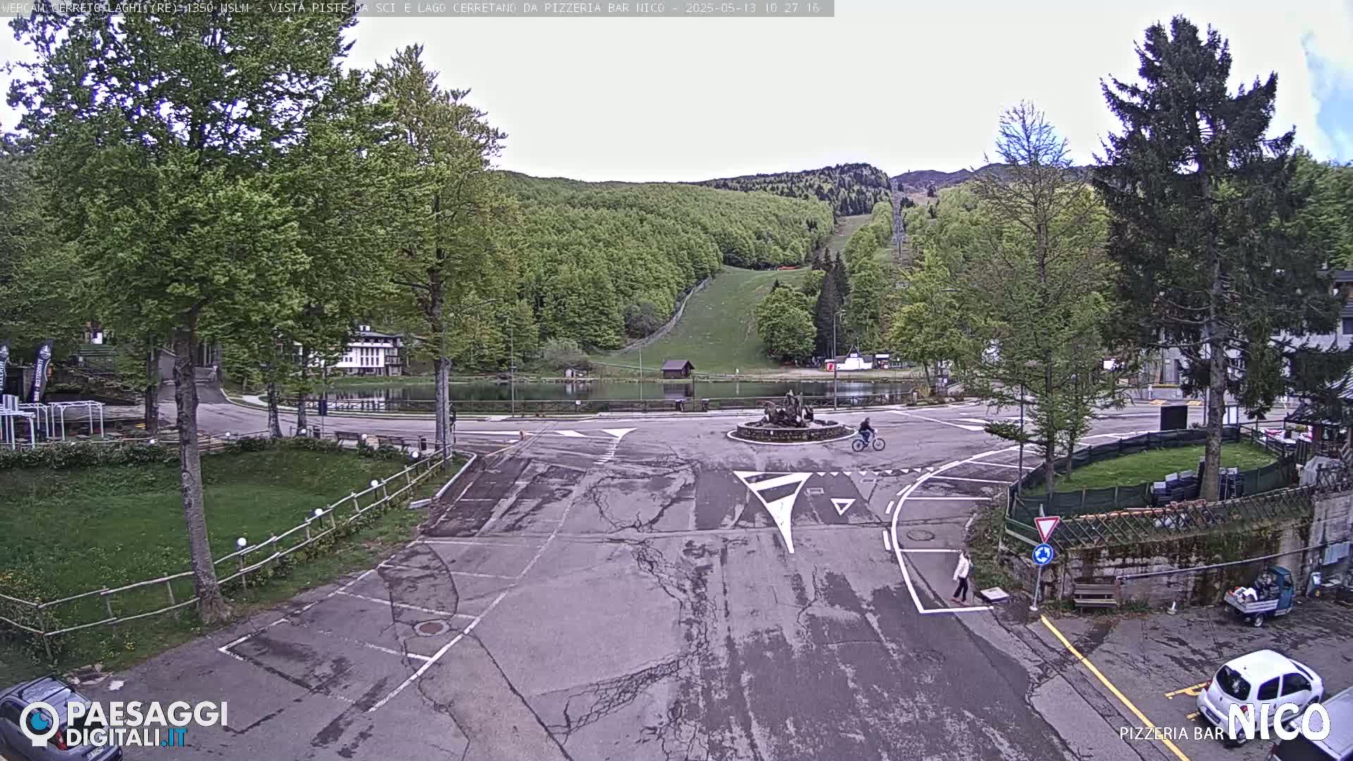A mostly overcast view of a parking lot and small lake, surrounded by green hills and trees, with a person riding a bicycle.