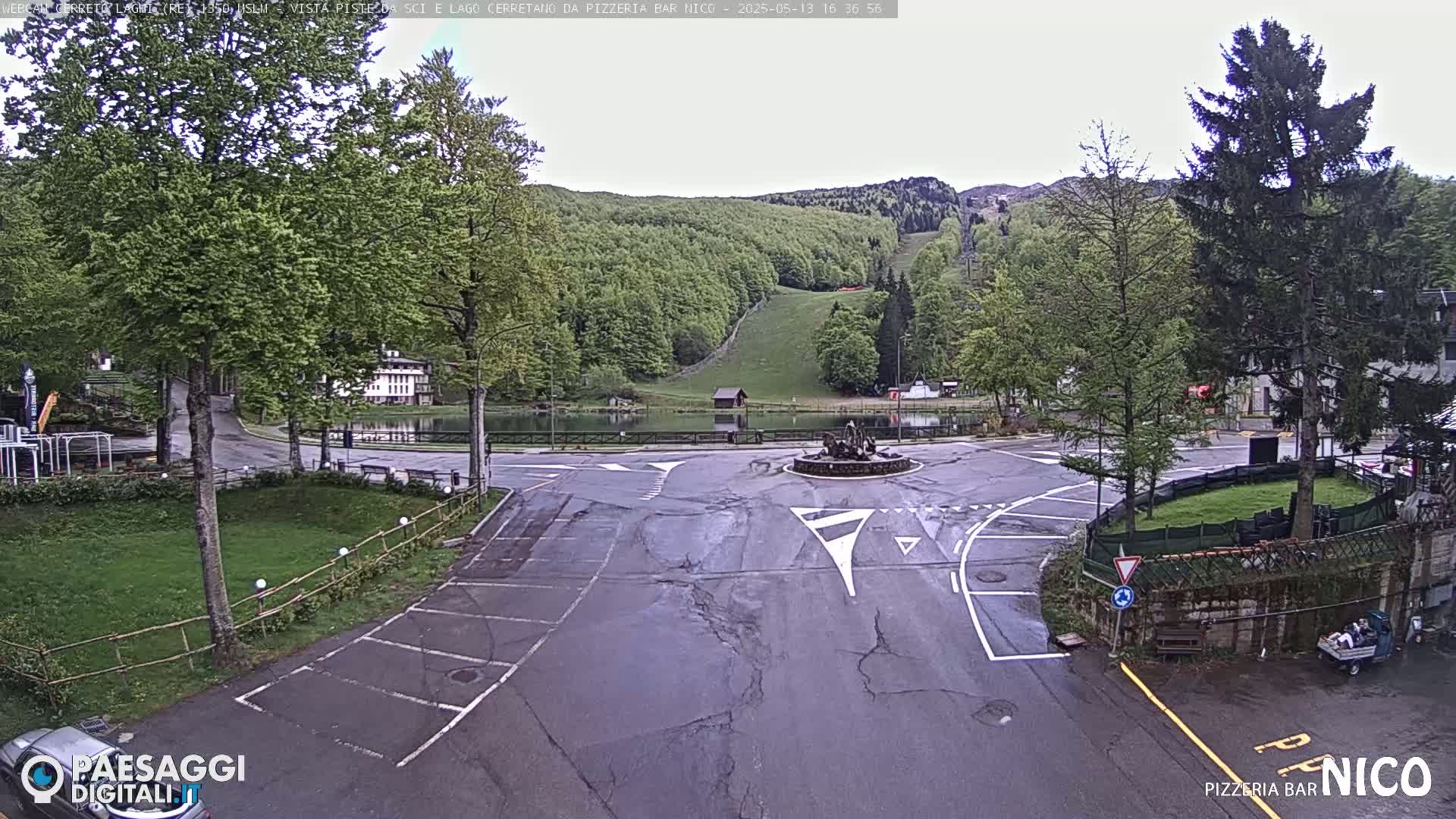 A wet roundabout near a small lake and green hills is surrounded by trees, buildings, and parking areas.