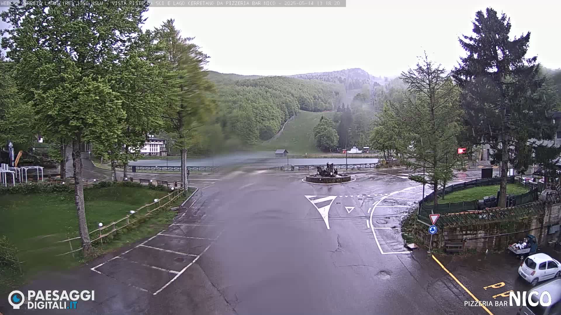 A wet, windy scene shows a parking lot and roadway leading to a lake and a hillside covered in trees and a ski slope.