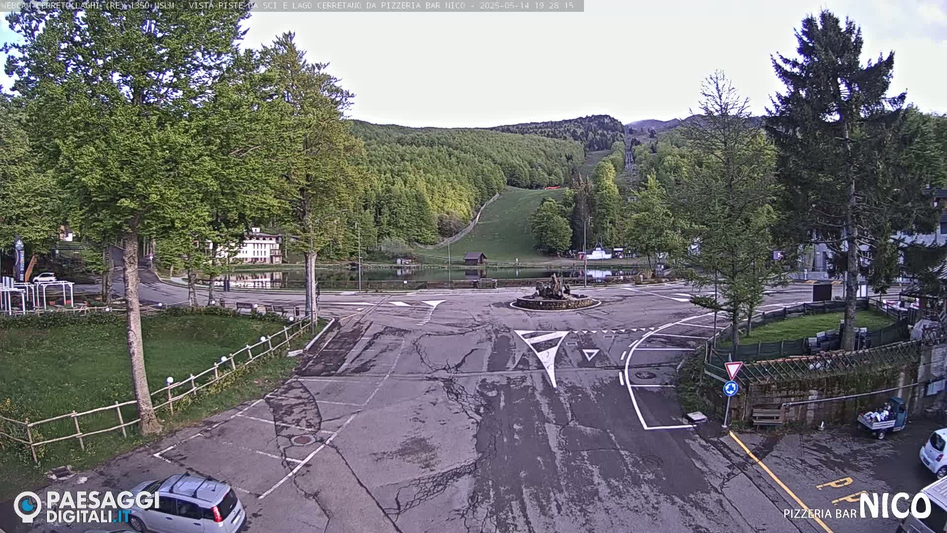 A mostly empty parking lot and roundabout are situated in front of a small town nestled at the base of a green, tree-covered hill.