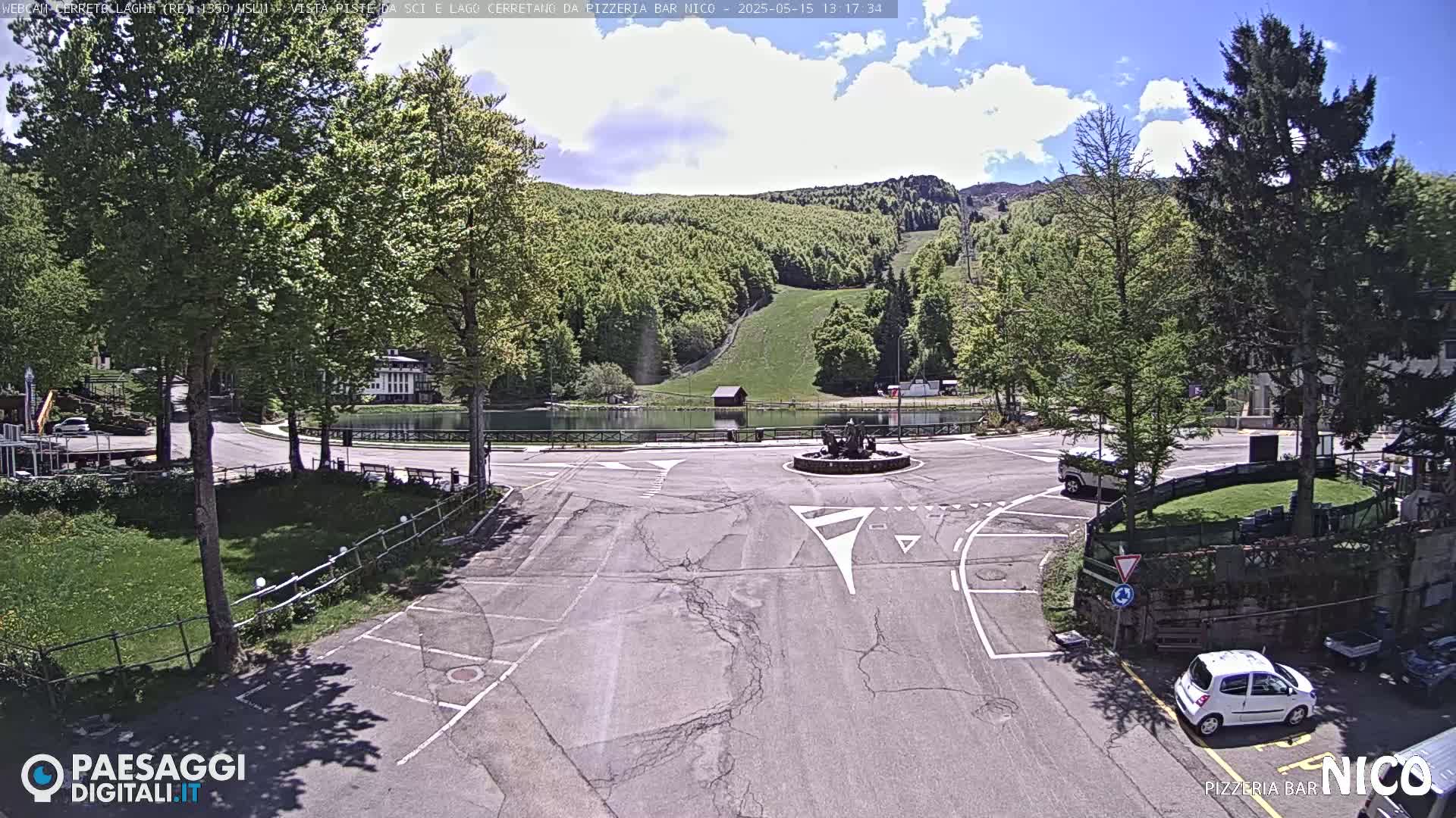 A mostly sunny day shows a parking lot and roundabout in front of a small pond, with lush green hills and trees in the background.