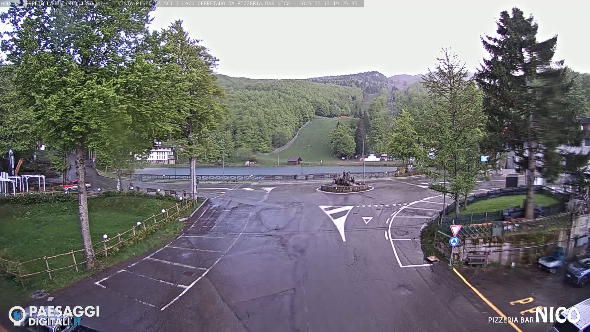 A wet, overcast day reveals a roundabout in a parking lot next to a tranquil lake and a hillside dotted with lush green trees.