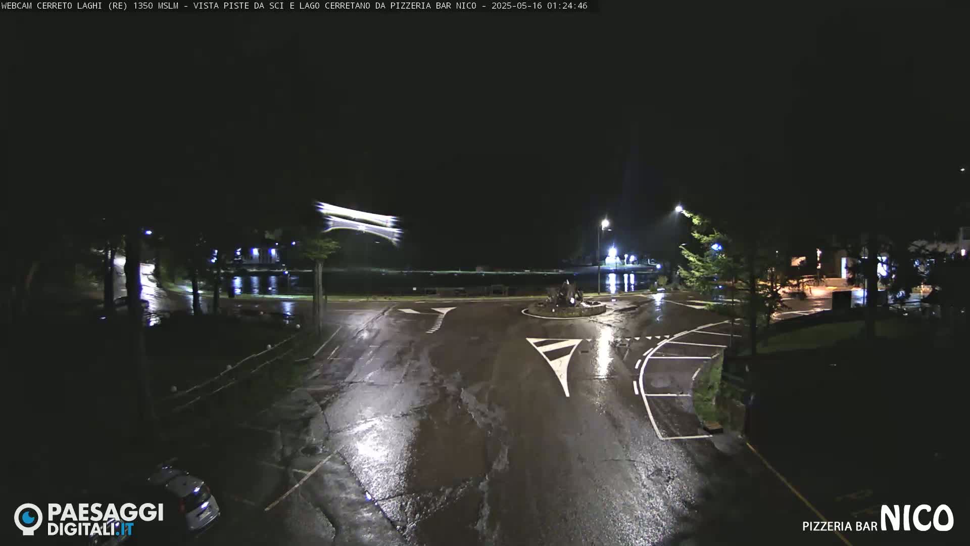 A wet, nighttime scene shows a roundabout and parking lot with a few cars, illuminated by streetlights.