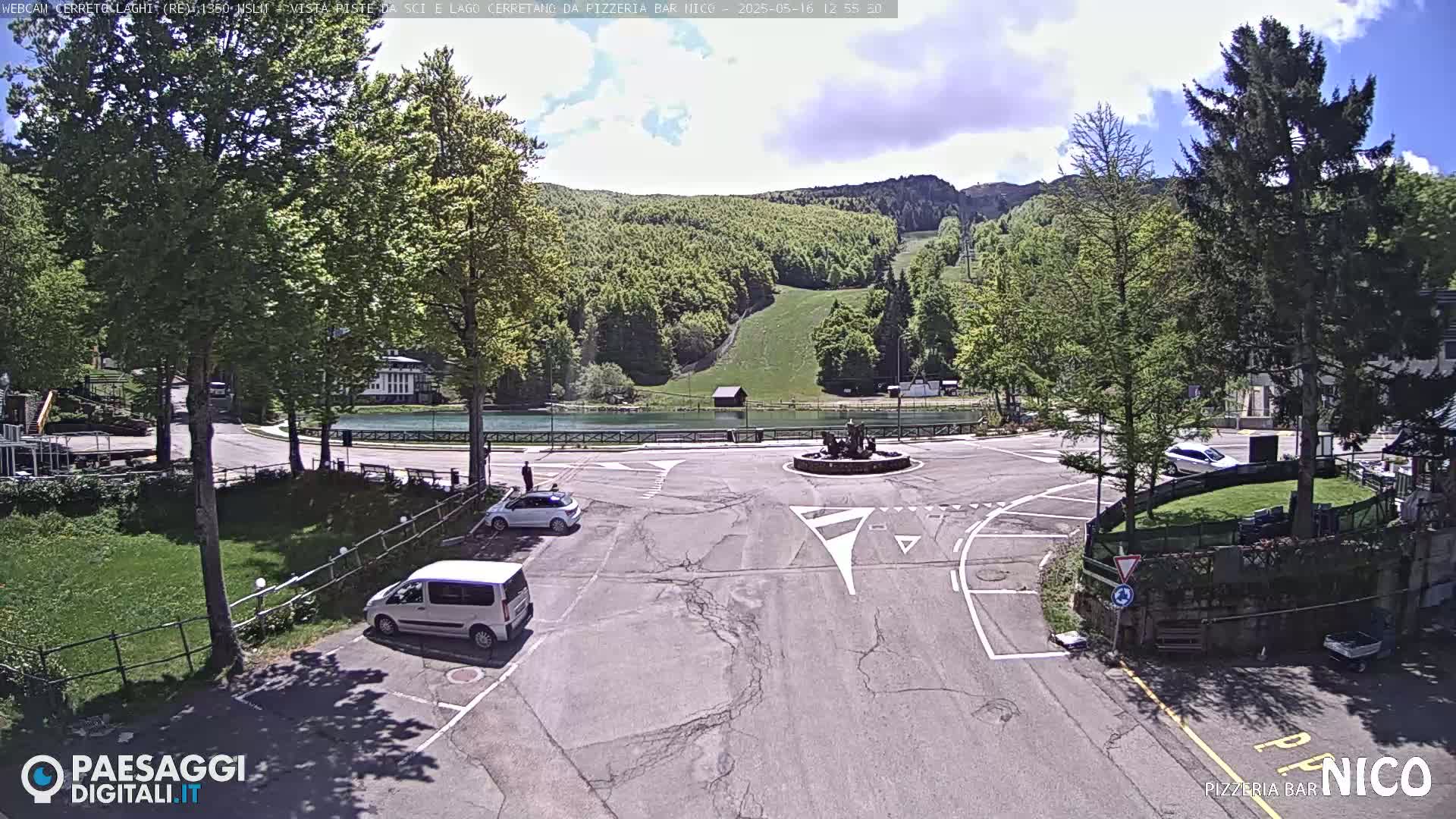 A sunny parking lot overlooks a small lake and a grassy hill with trees, under a partly cloudy sky.
