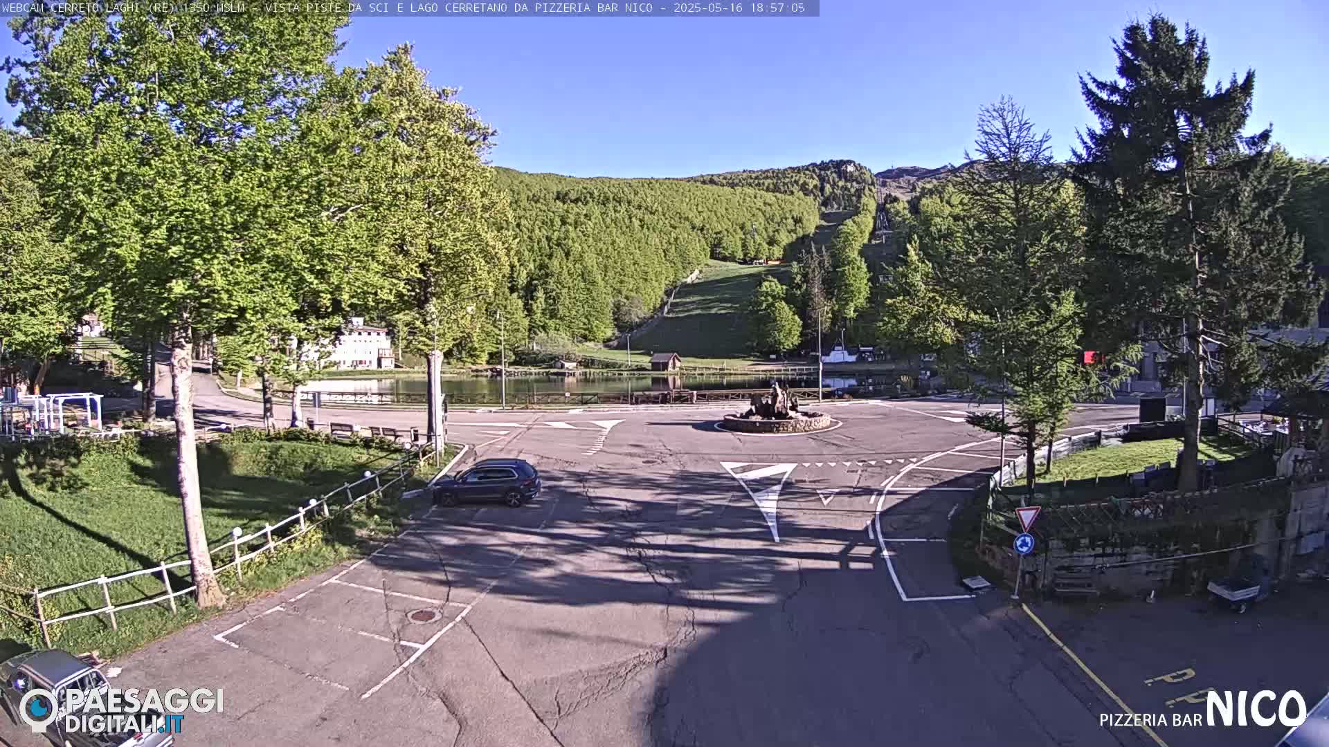 A sunny, outdoor view of a small town square with a pond, a fountain, and a car parked in a nearby lot, nestled against a backdrop of a mountainside forest.