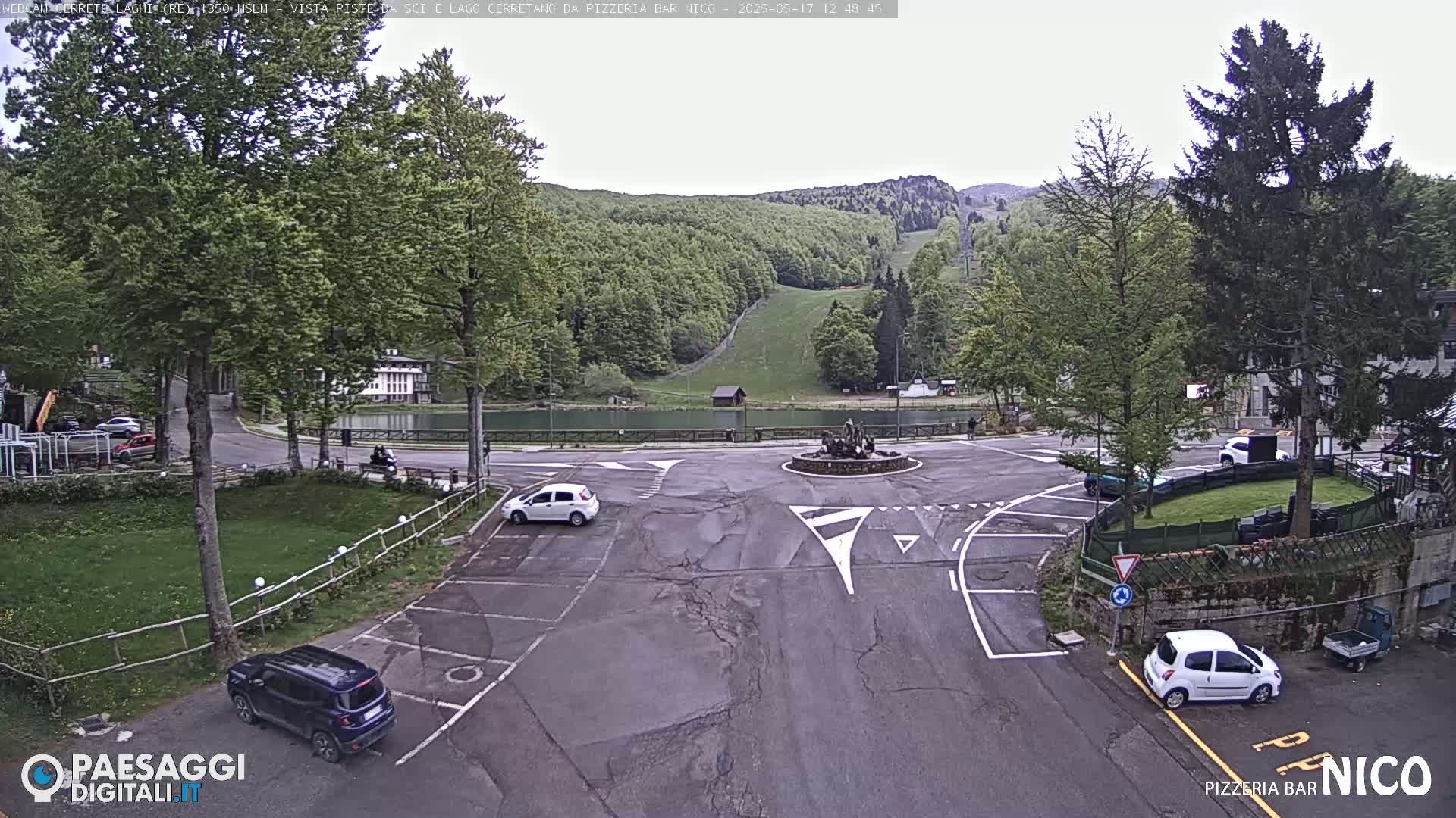 A parking lot and roundabout overlook a calm lake and a hillside with green trees and a ski slope under an overcast sky.