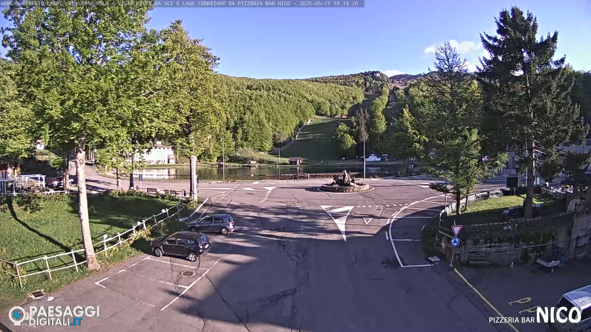 A sunny view of a parking lot, small pond, and roundabout, situated in a mountainous area with lush green trees and a ski slope visible in the background.