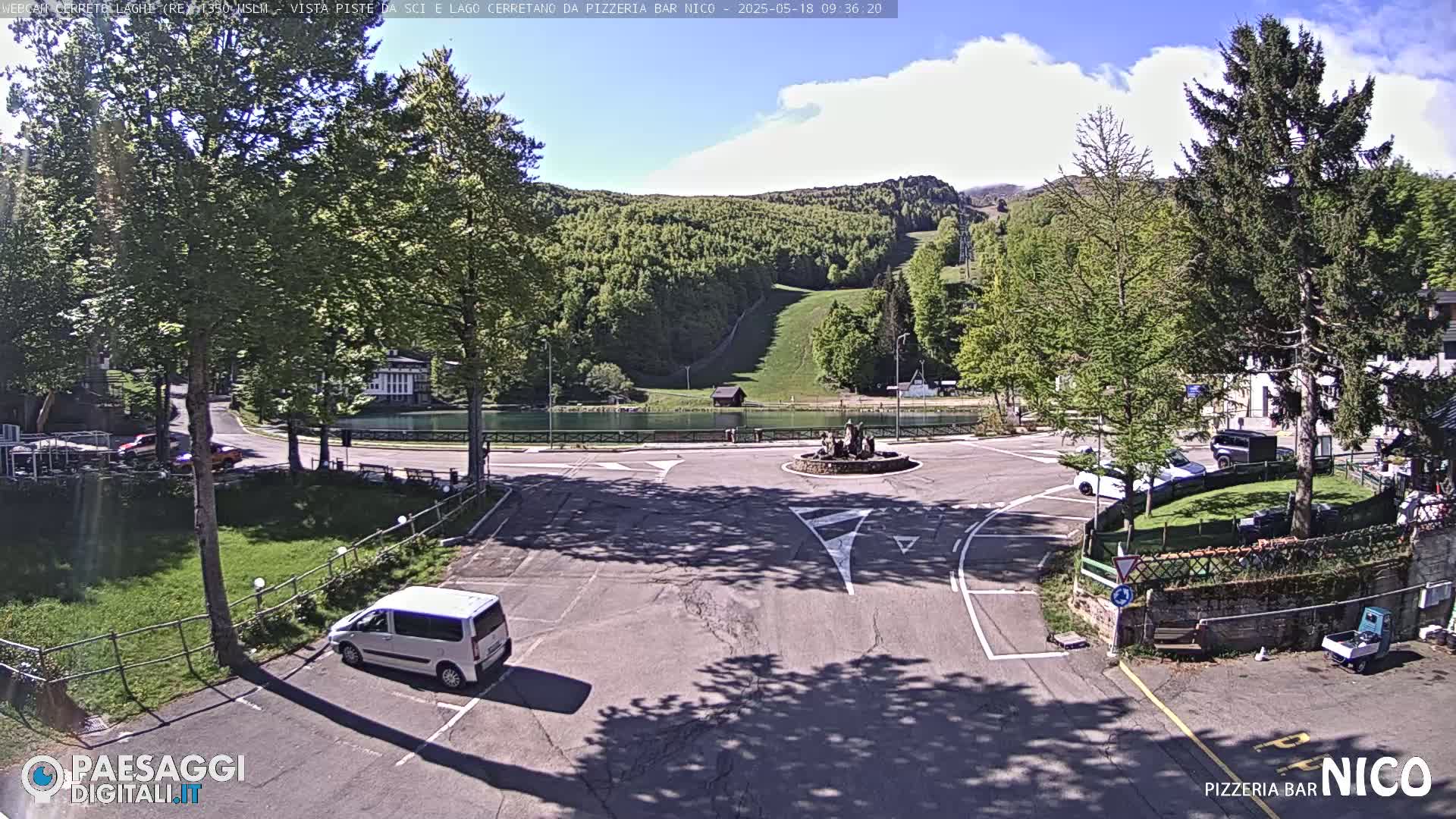 A sunny day at a mountainside roundabout with a small lake and ski slope visible in the background, featuring a white van parked in a lot, and several cars visible further away.