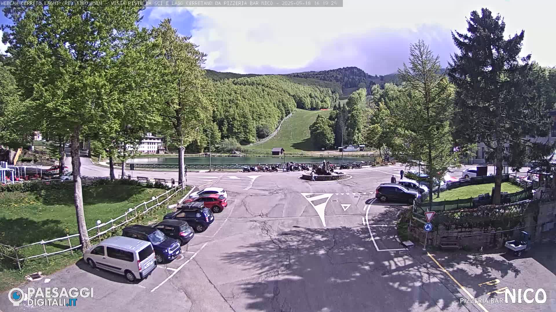 A sunny day shows a parking lot with several cars, a small lake, and a hillside with a ski slope in the background.