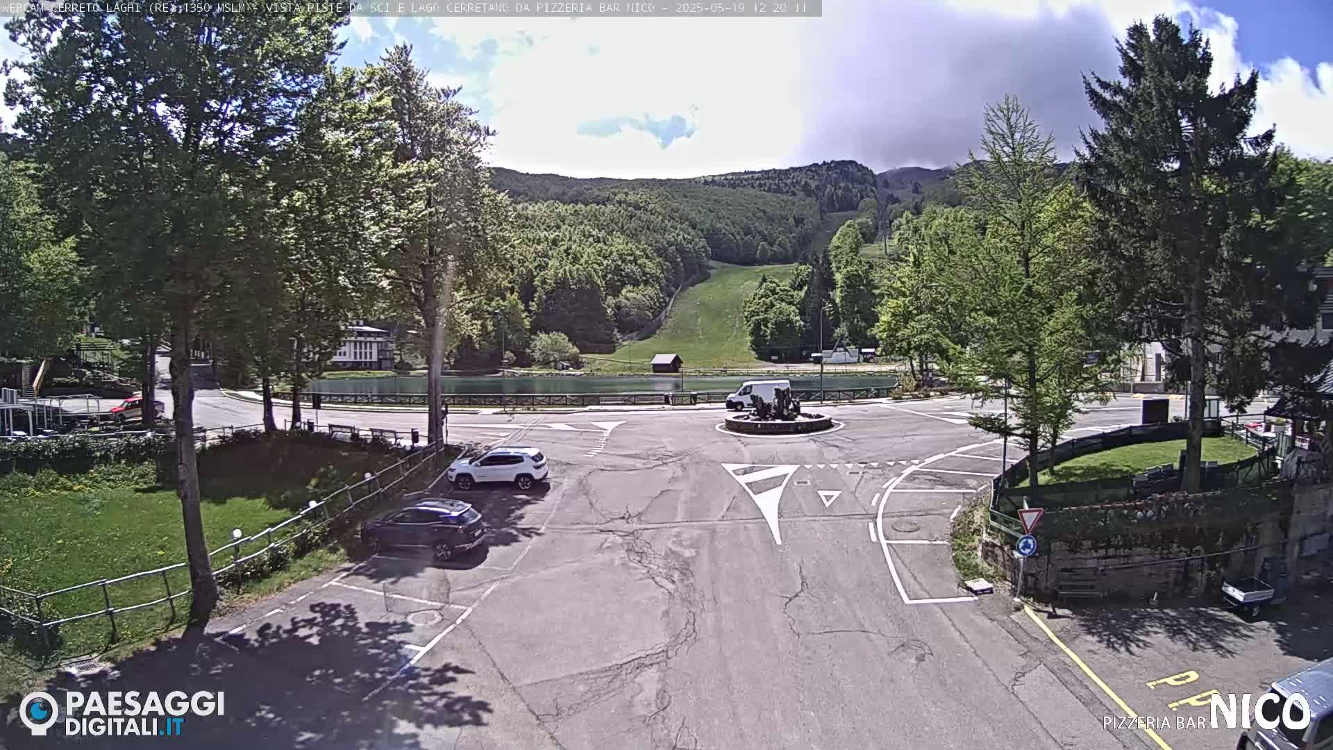 A mostly sunny day shows a parking lot and roadway near a small lake and a ski slope in a mountainous region.