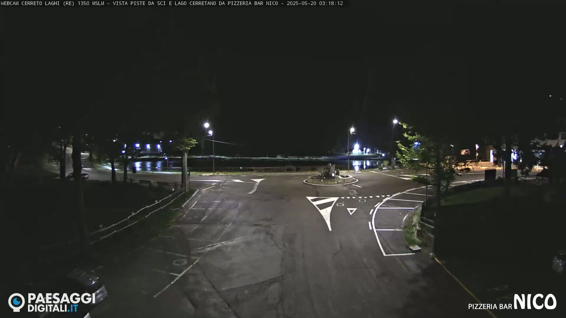 A nighttime view of a road intersection and parking lot near a body of water, illuminated by streetlights.