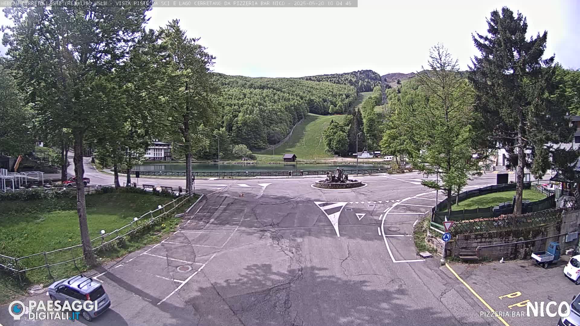 A parking lot and roundabout are situated before a small lake, beyond which are green hills with a ski lift visible in the distance, under a mostly clear sky.