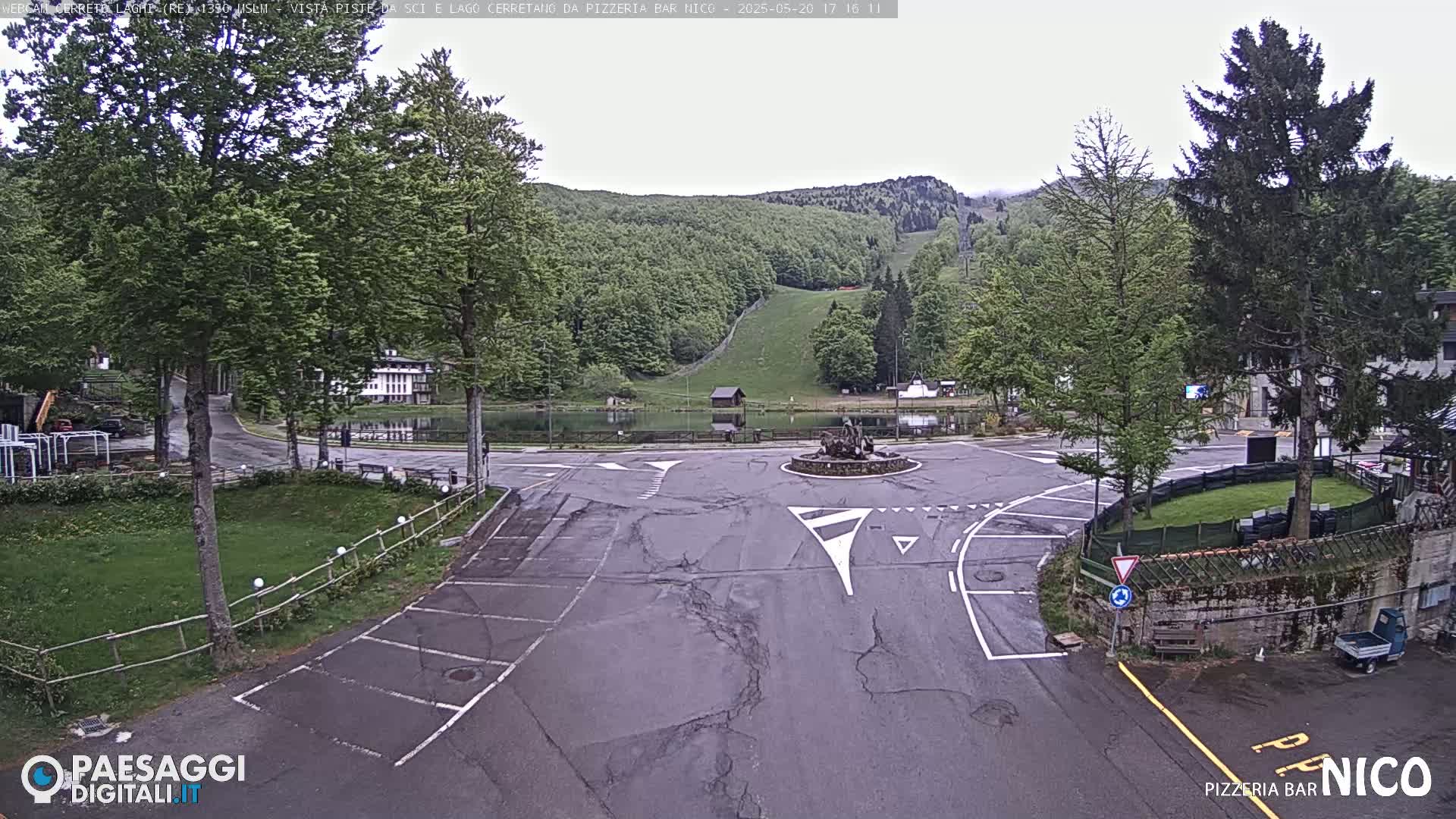 A wet asphalt road and parking area curve around a small pond and grassy area, with a hillside covered in lush green trees and a ski slope visible in the background under an overcast sky.
