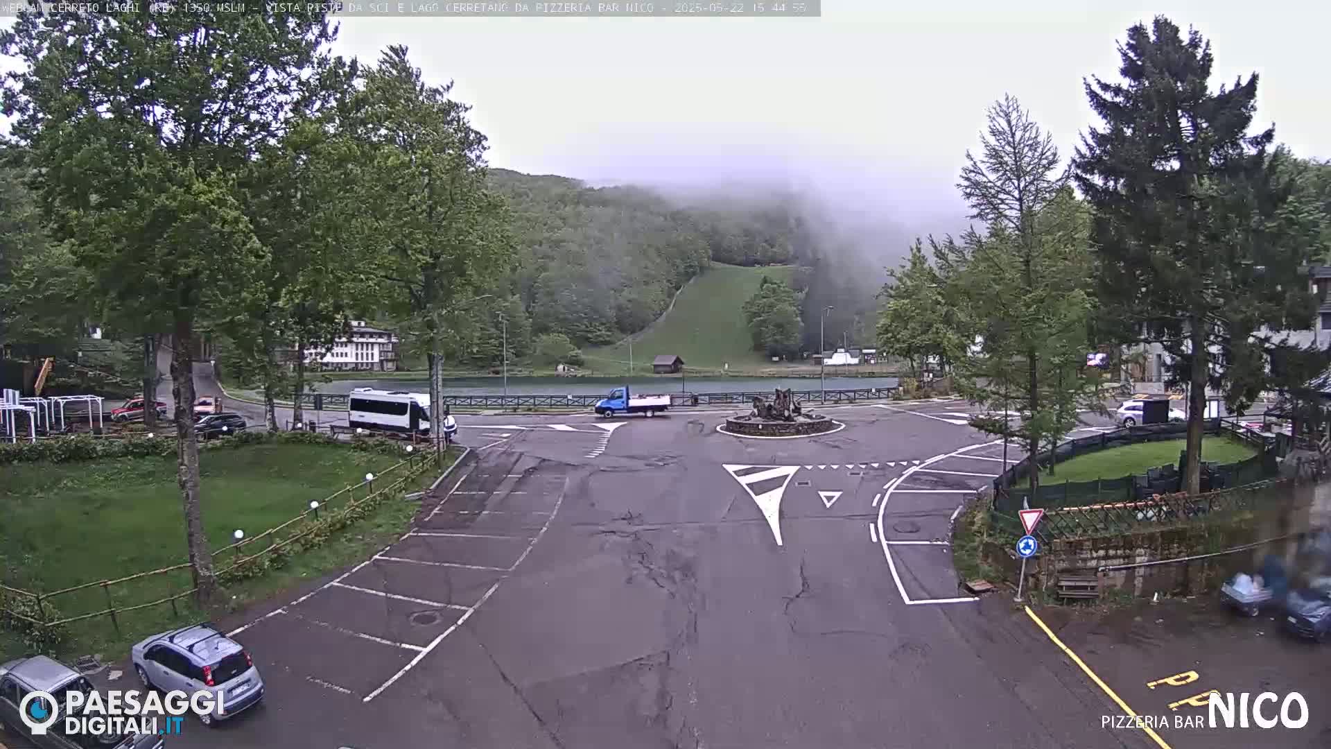 A foggy mountainside scene shows a roundabout with several vehicles parked and driving, surrounded by lush green trees and a small body of water.