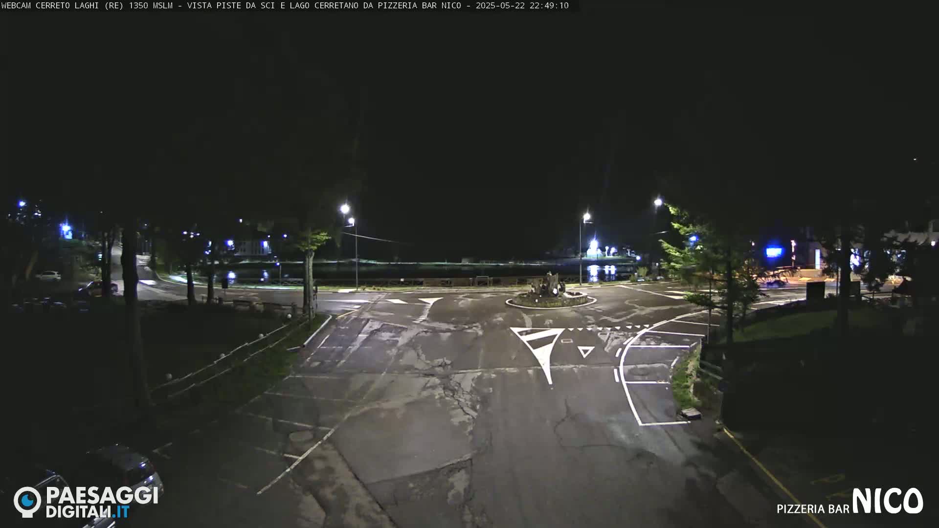 A nighttime view of a paved intersection and roundabout near a body of water, illuminated by streetlights.