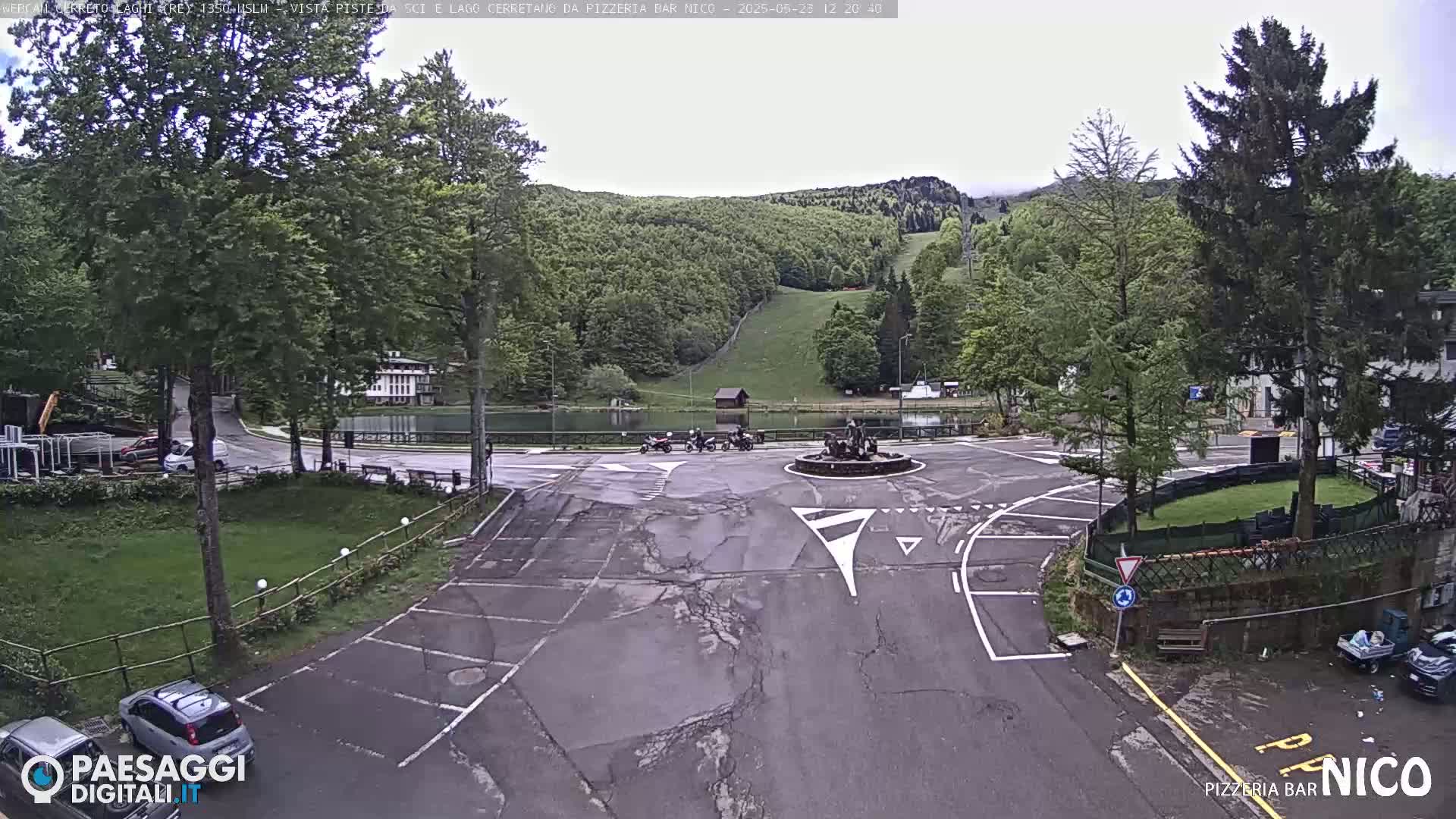 An overcast day reveals a parking lot and roundabout in a village setting, with a small lake and a grassy hillside visible in the background.