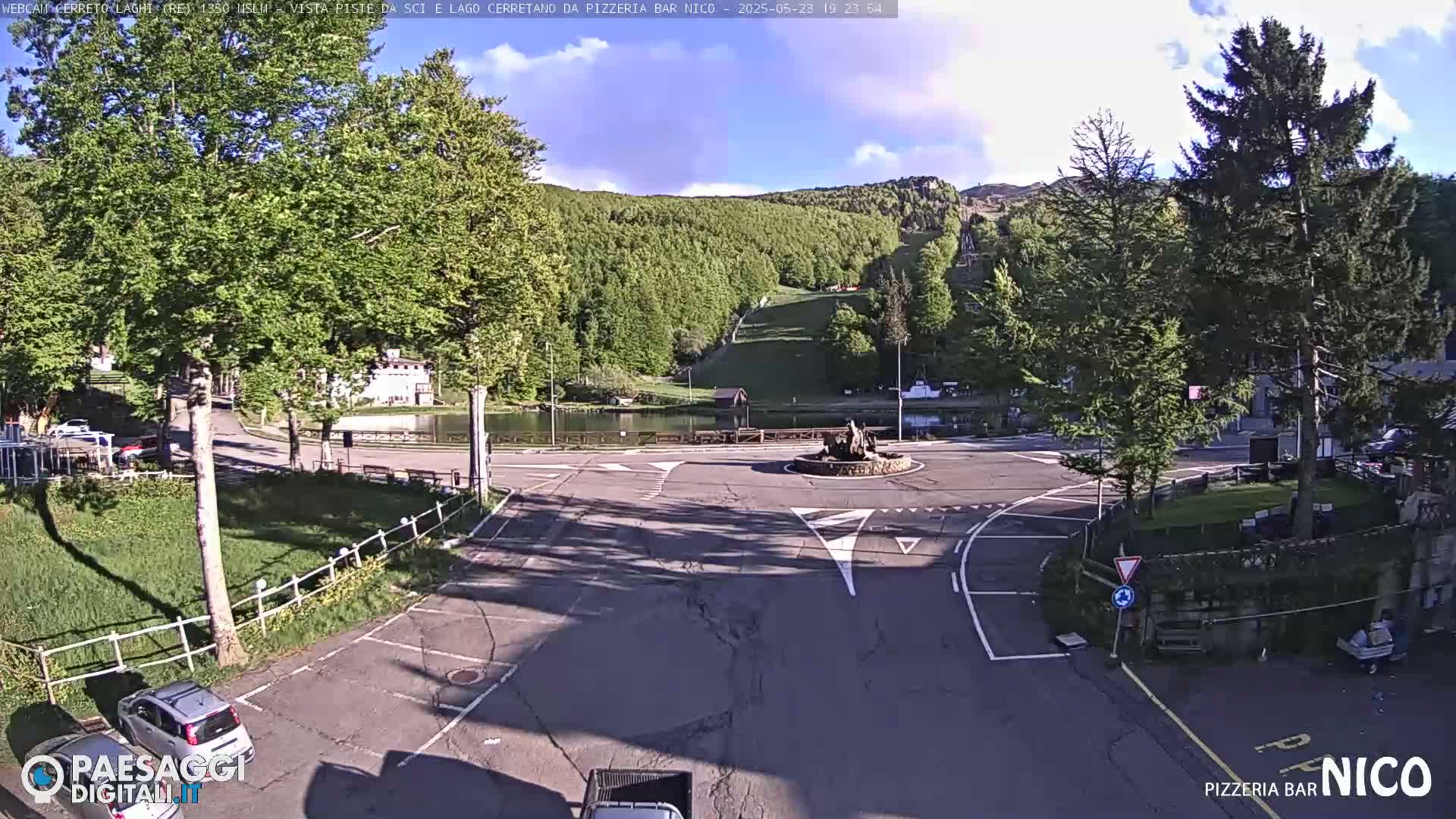 A mostly sunny view of a paved roundabout with a small pond and trees, surrounded by buildings and a parking lot, next to a forested hillside.