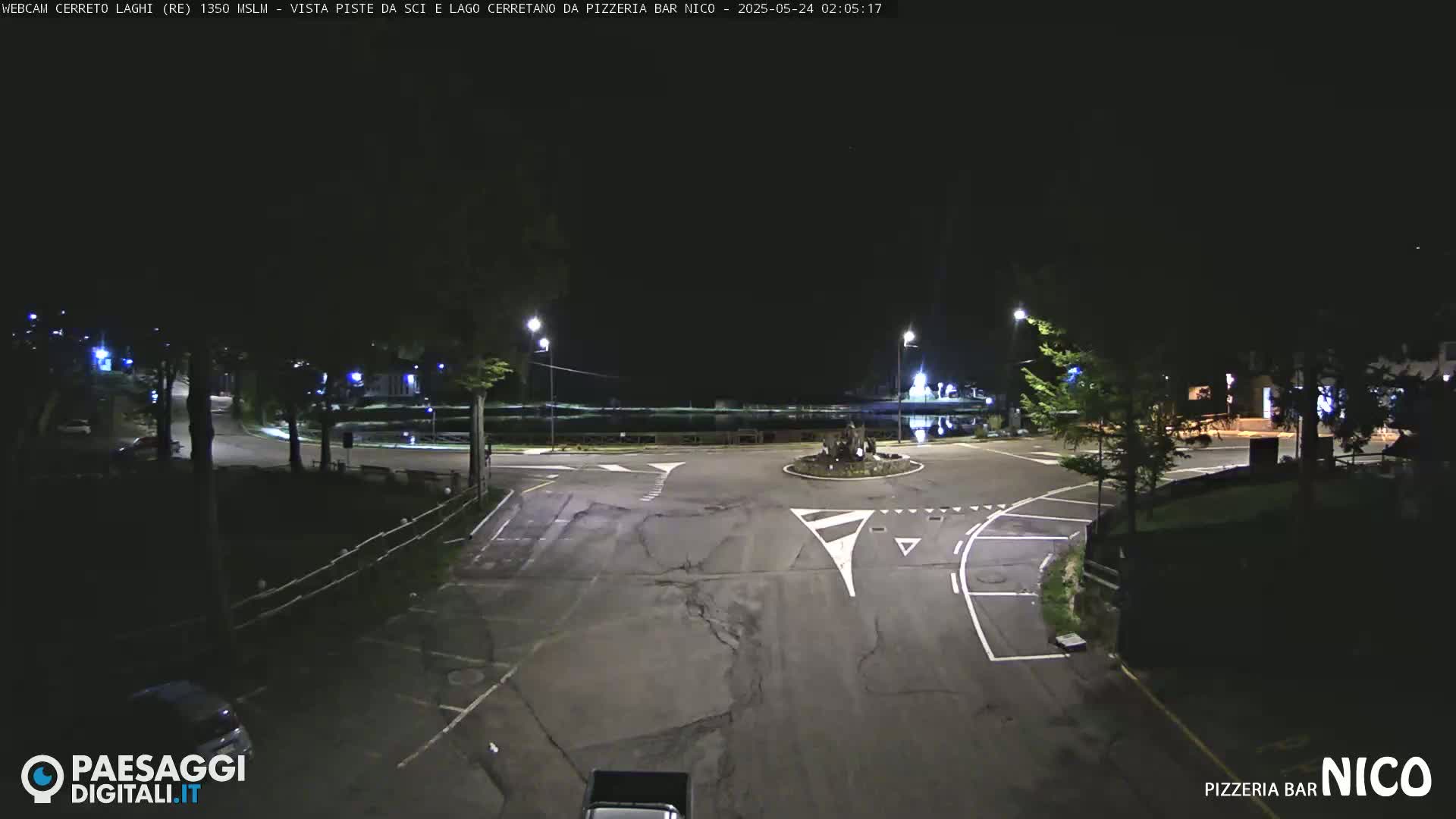A dark, nighttime view of an empty road intersection with some streetlights and trees surrounding it.