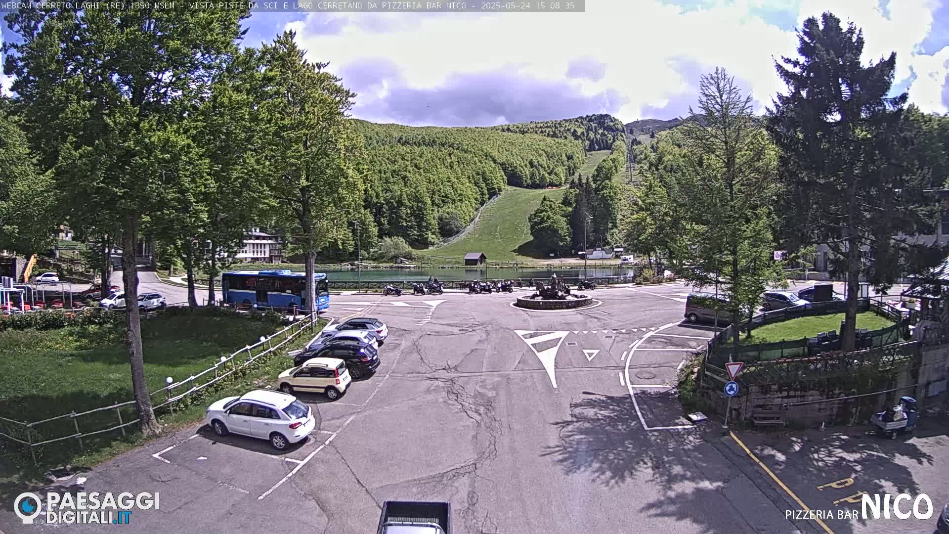 A mostly sunny day shows a parking lot with cars and a bus near a roundabout, overlooking a lake, a ski slope, and a green mountainside.