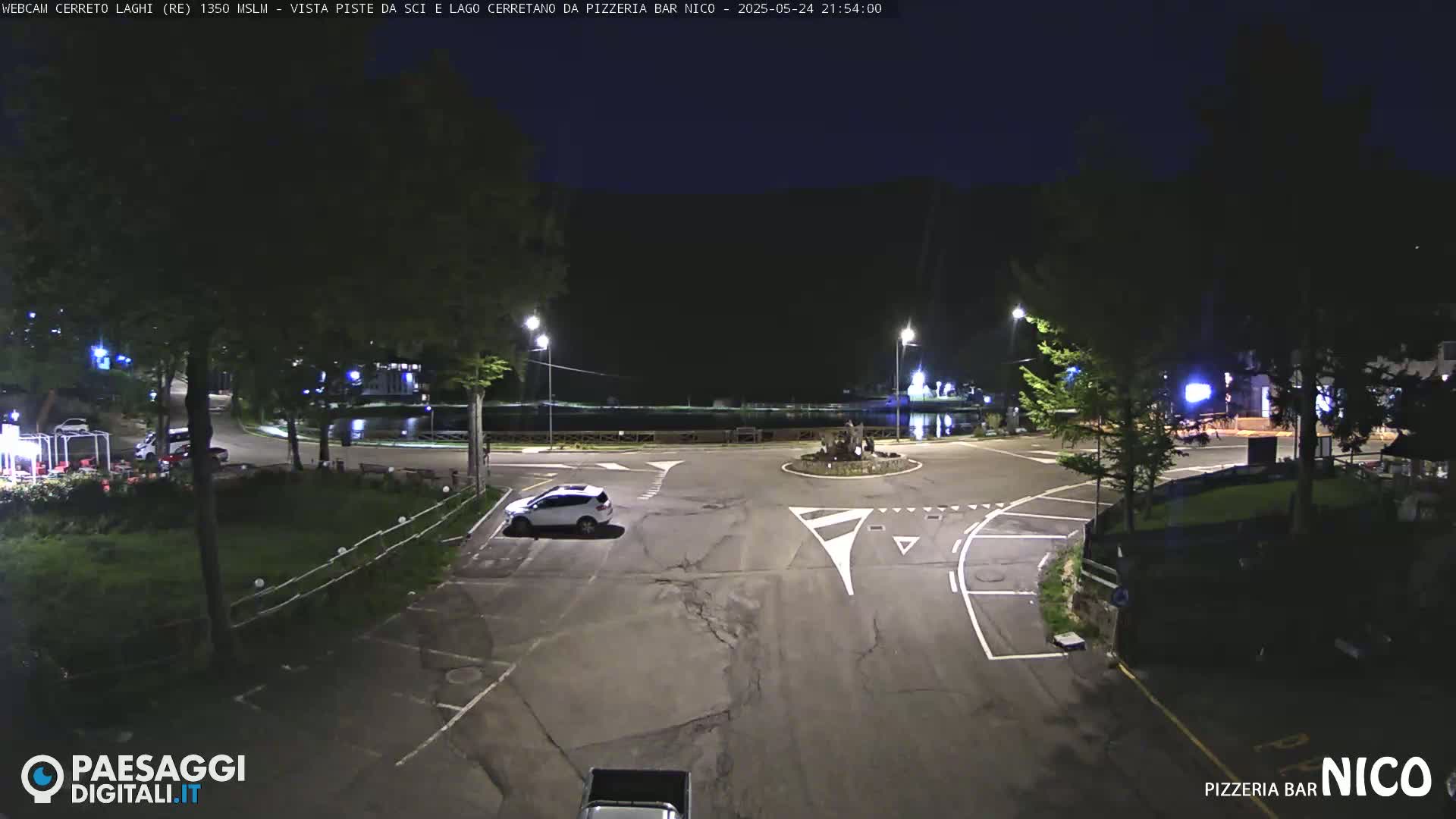 A nighttime view of a mostly empty parking lot and roundabout near a body of water, under a dark sky.
