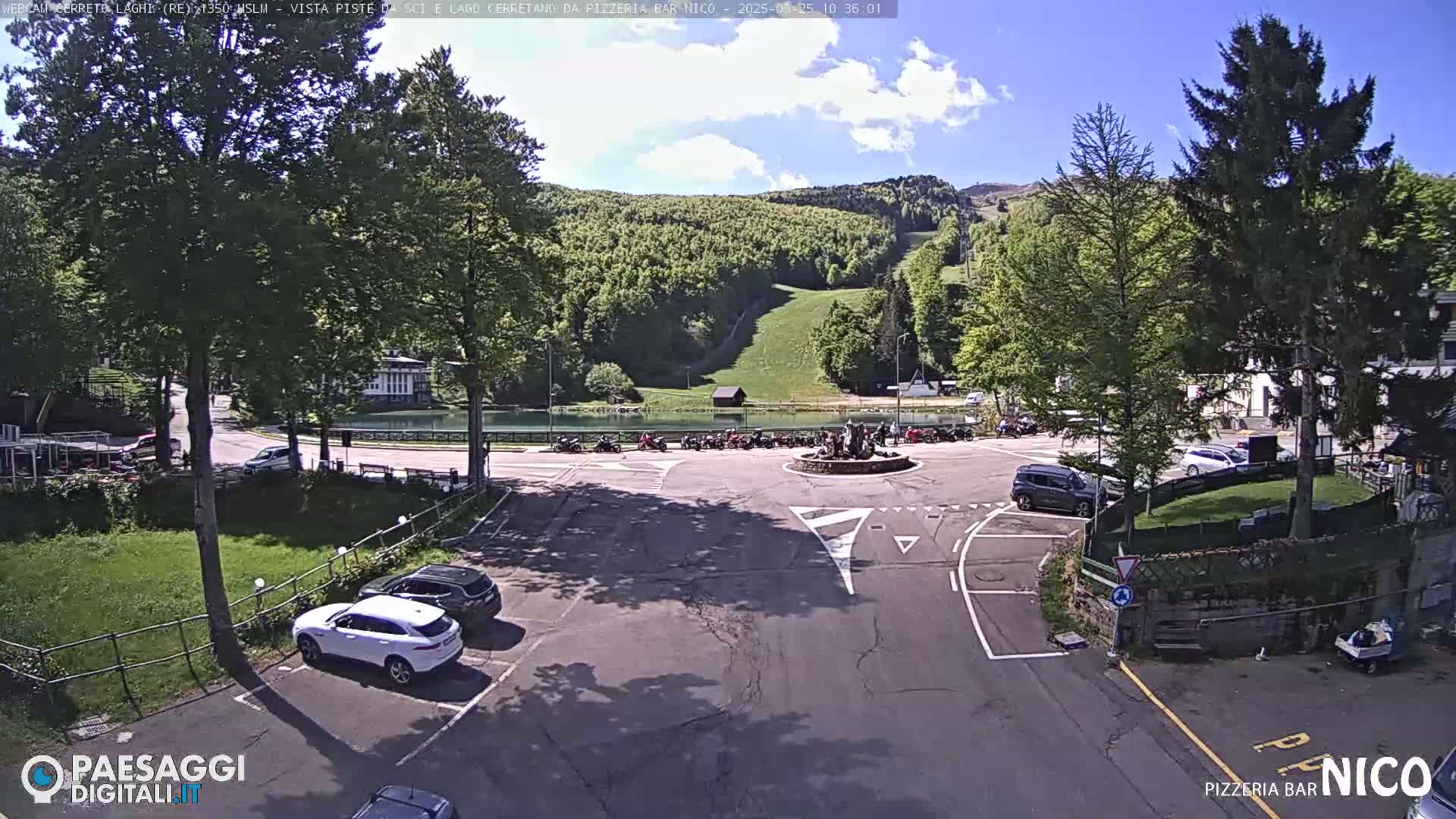 A sunny day reveals a parking lot, a small lake, and a grassy hillside with a ski slope in the background.