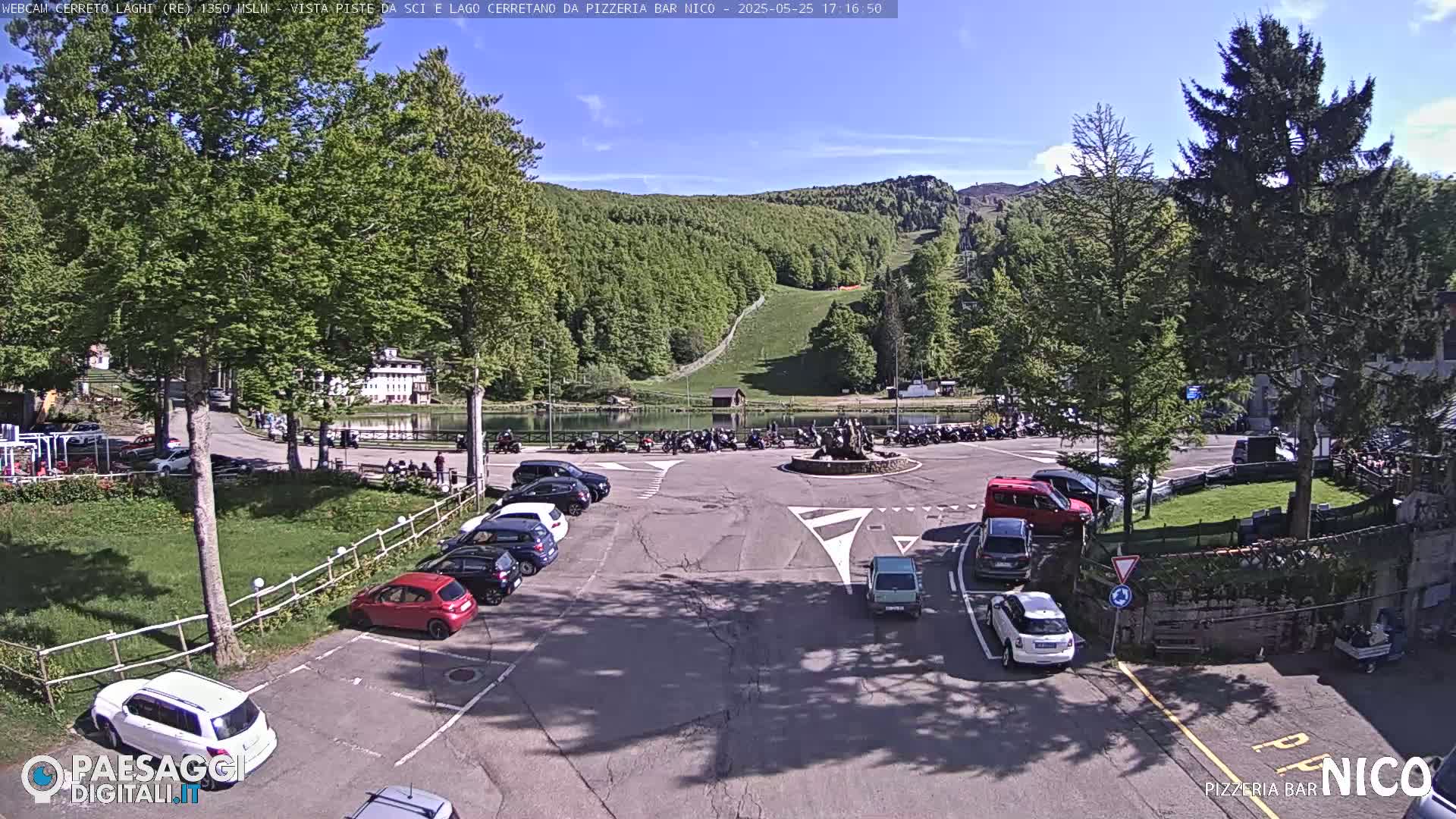 A parking lot with several cars is situated near a small lake and a grassy hill with a ski lift in the background, under a sunny sky.