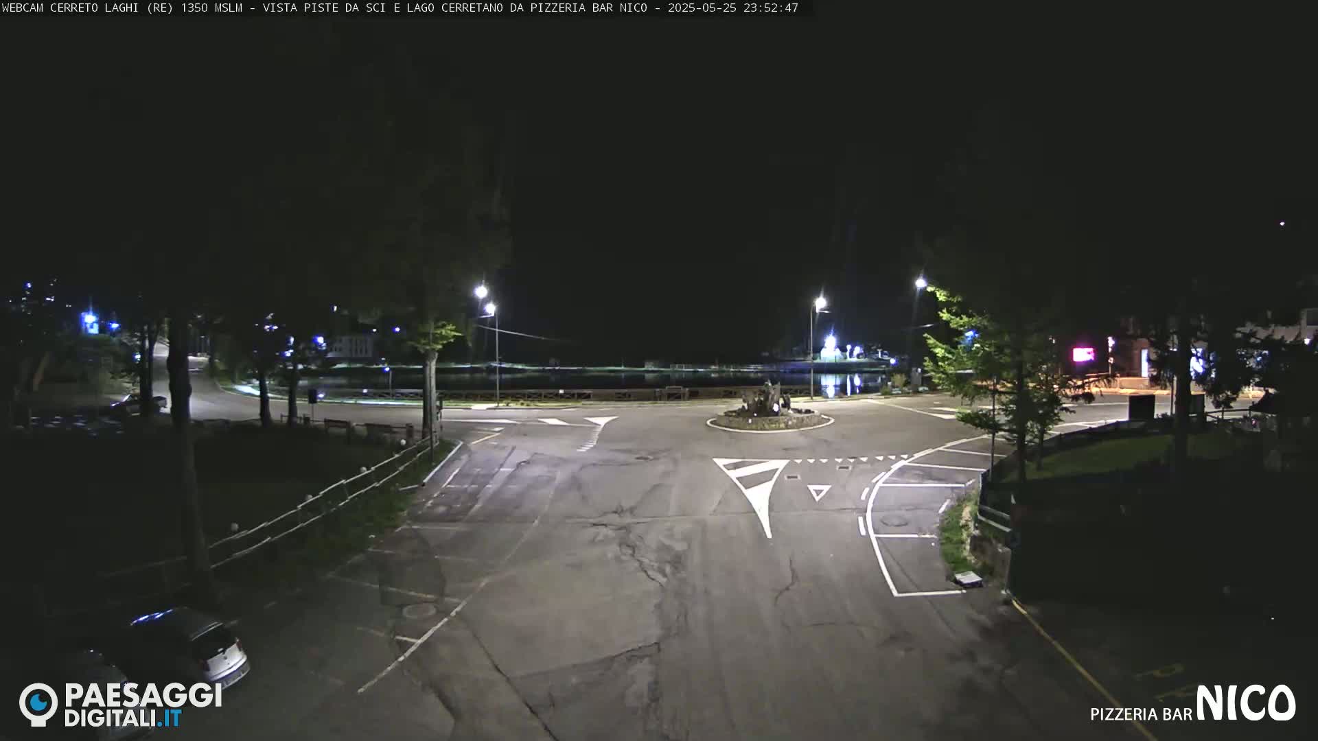 A nighttime view of a dark, empty roundabout and parking lot, illuminated by streetlights.