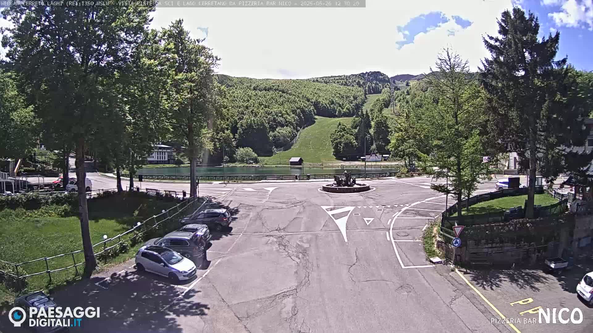 A sunny day shows a parking lot with several cars, a circular roadway around a fountain, and a small lake next to a grassy hillside with trees.